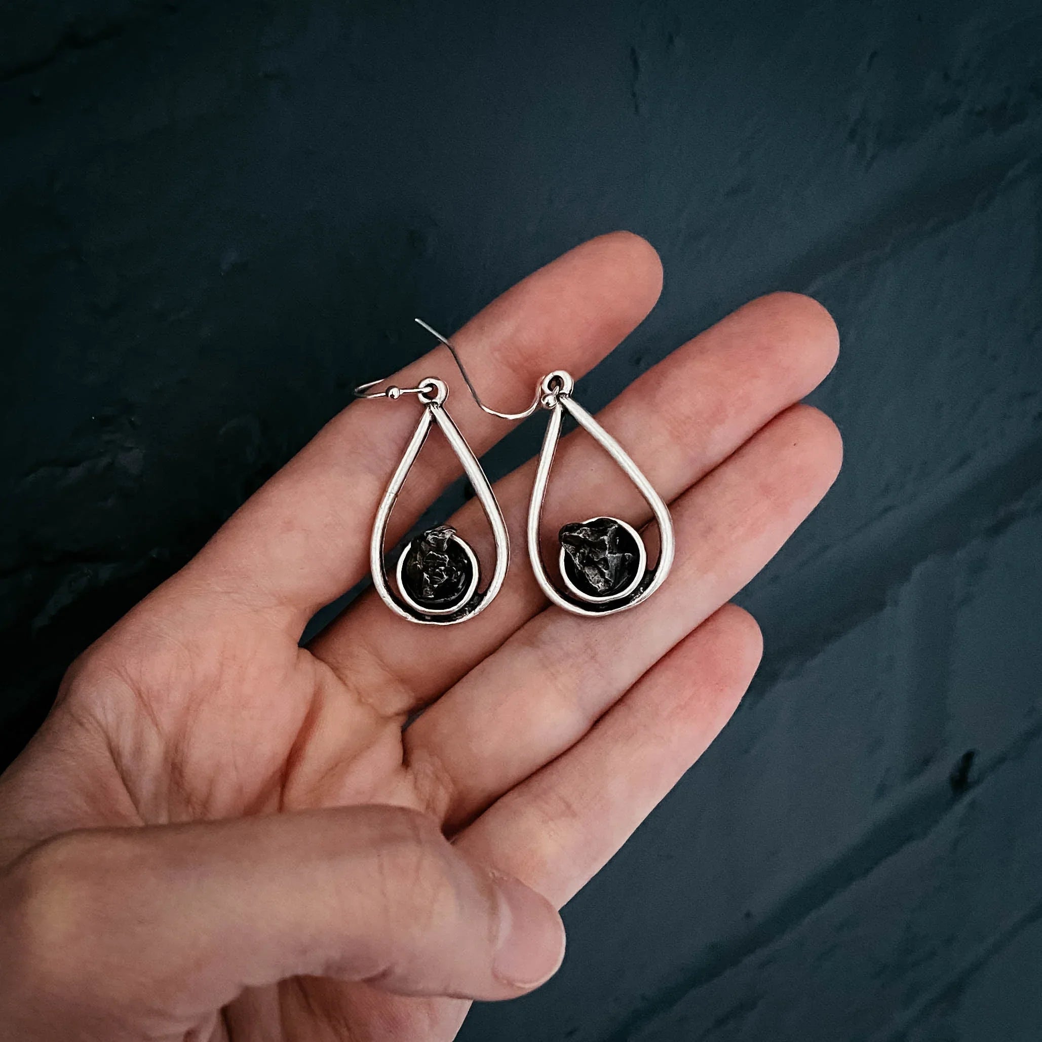 Hand holding silver teardrop earrings with black textured stone center on dark background