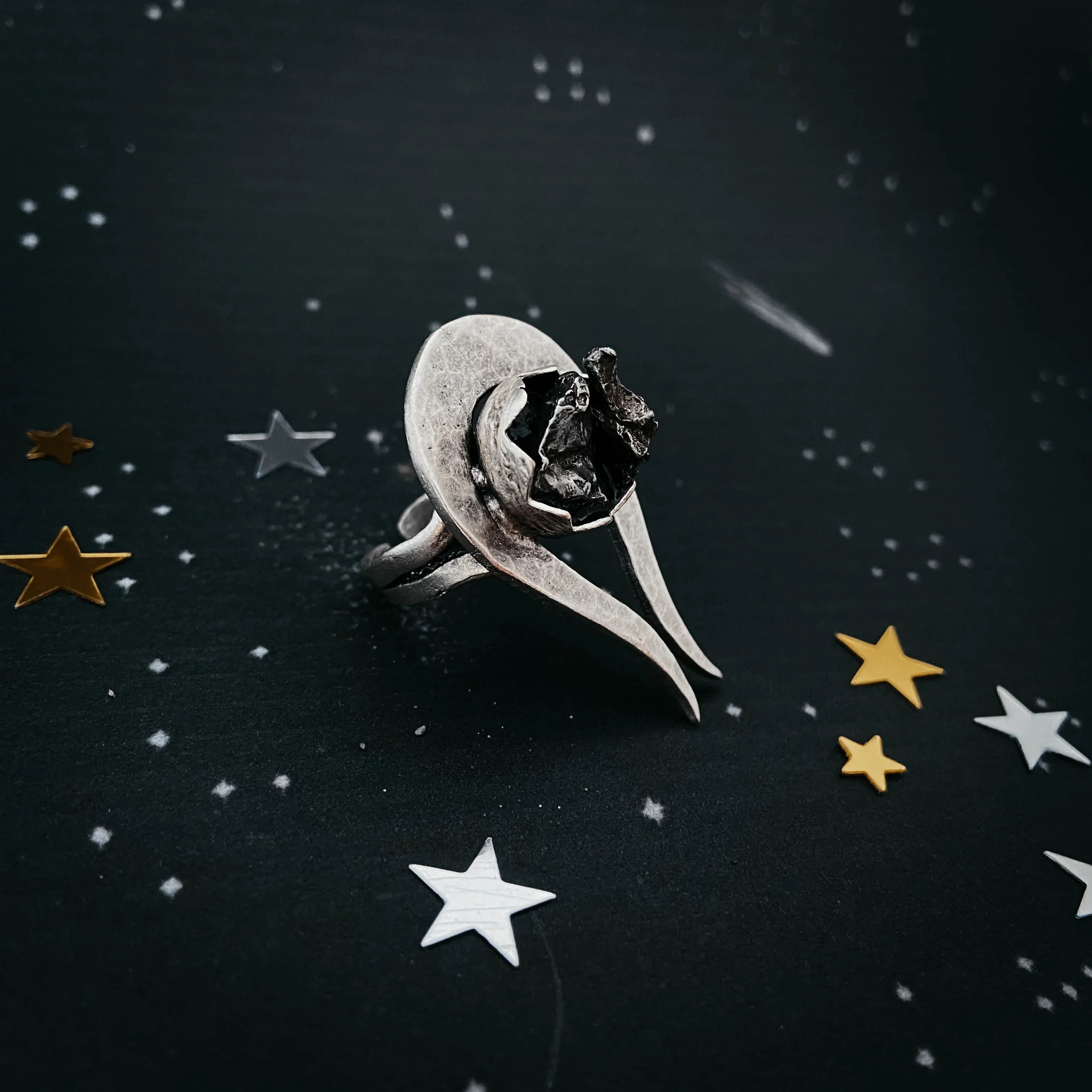 unique silver ring with irregular dark stone on starry black background with gold and silver star confetti