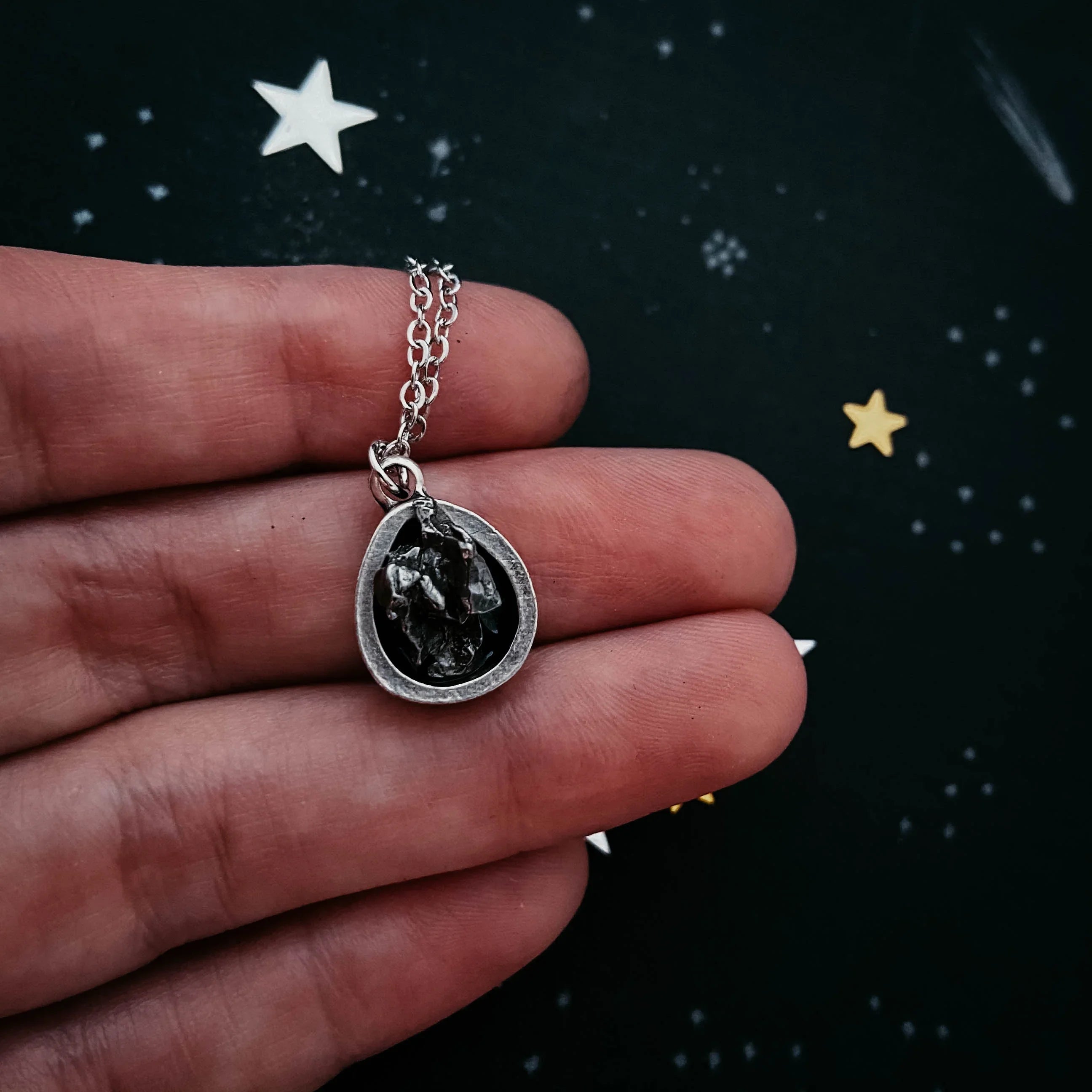 Hand holding a cosmic-themed silver pendant necklace with black stone on starry background