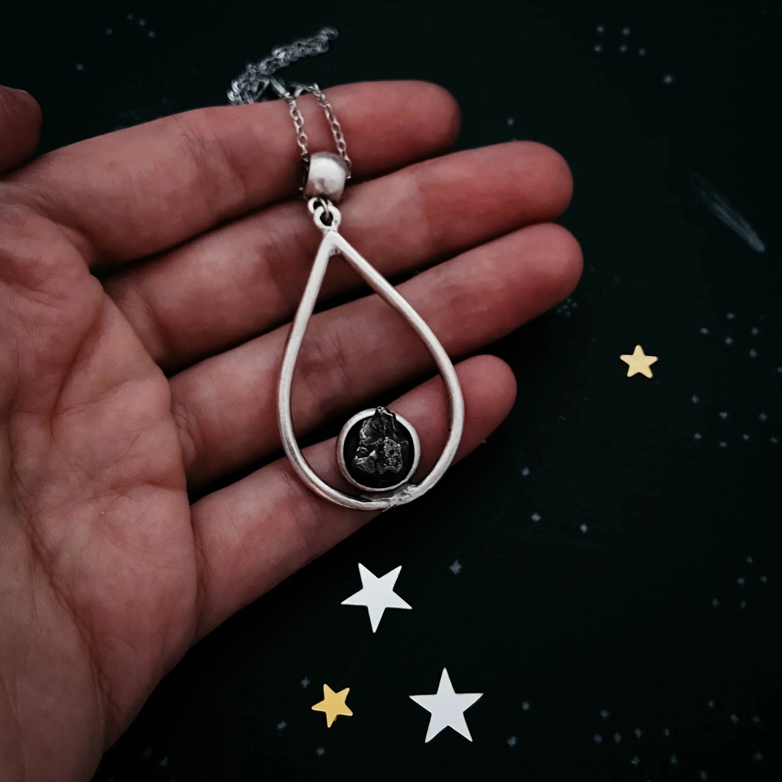 Hand holding silver teardrop pendant necklace with black stone against starry dark background