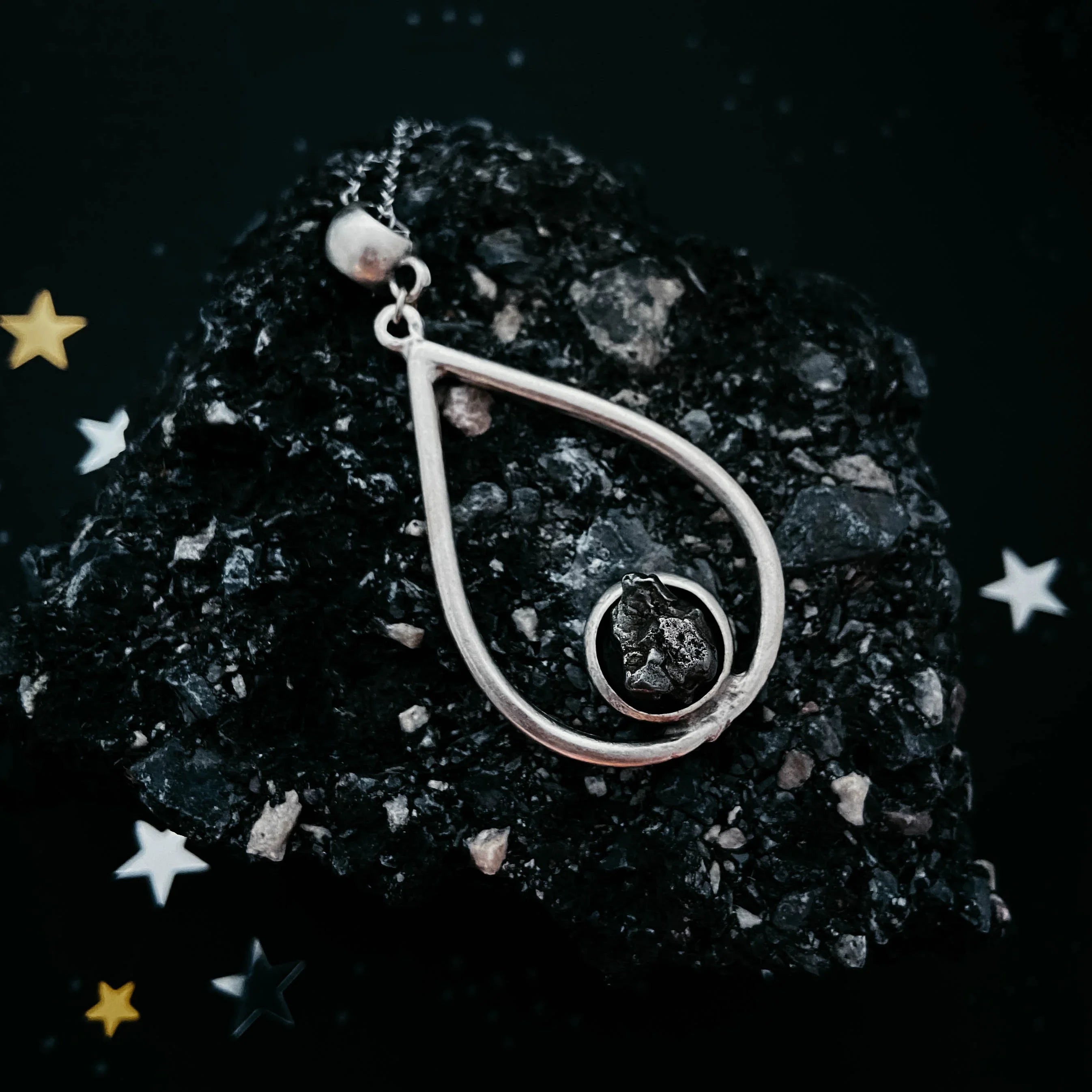 Silver teardrop pendant with a raw black stone on textured black rock with star confetti