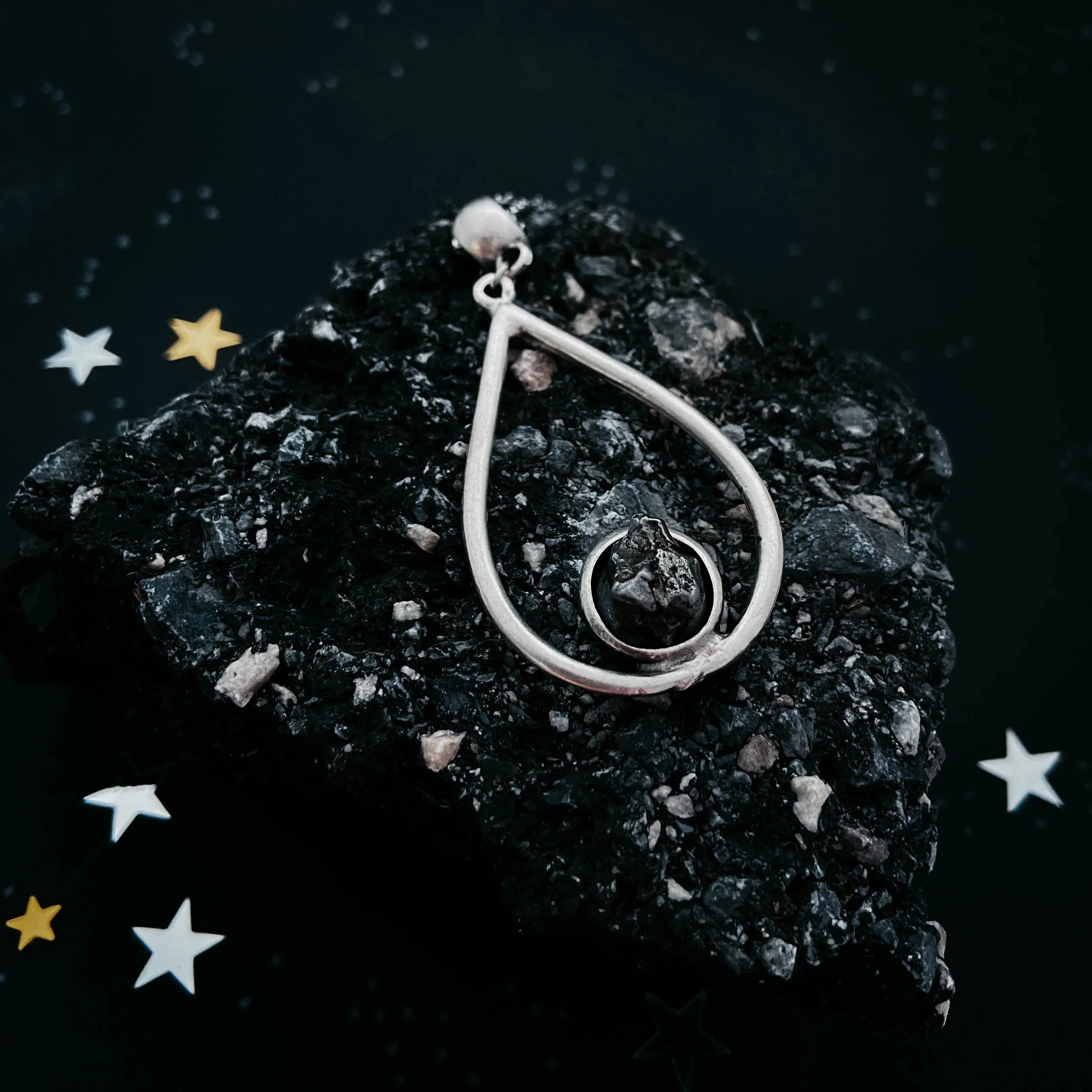 Silver teardrop pendant with a dark stone resting on a textured black rock with star-shaped confetti