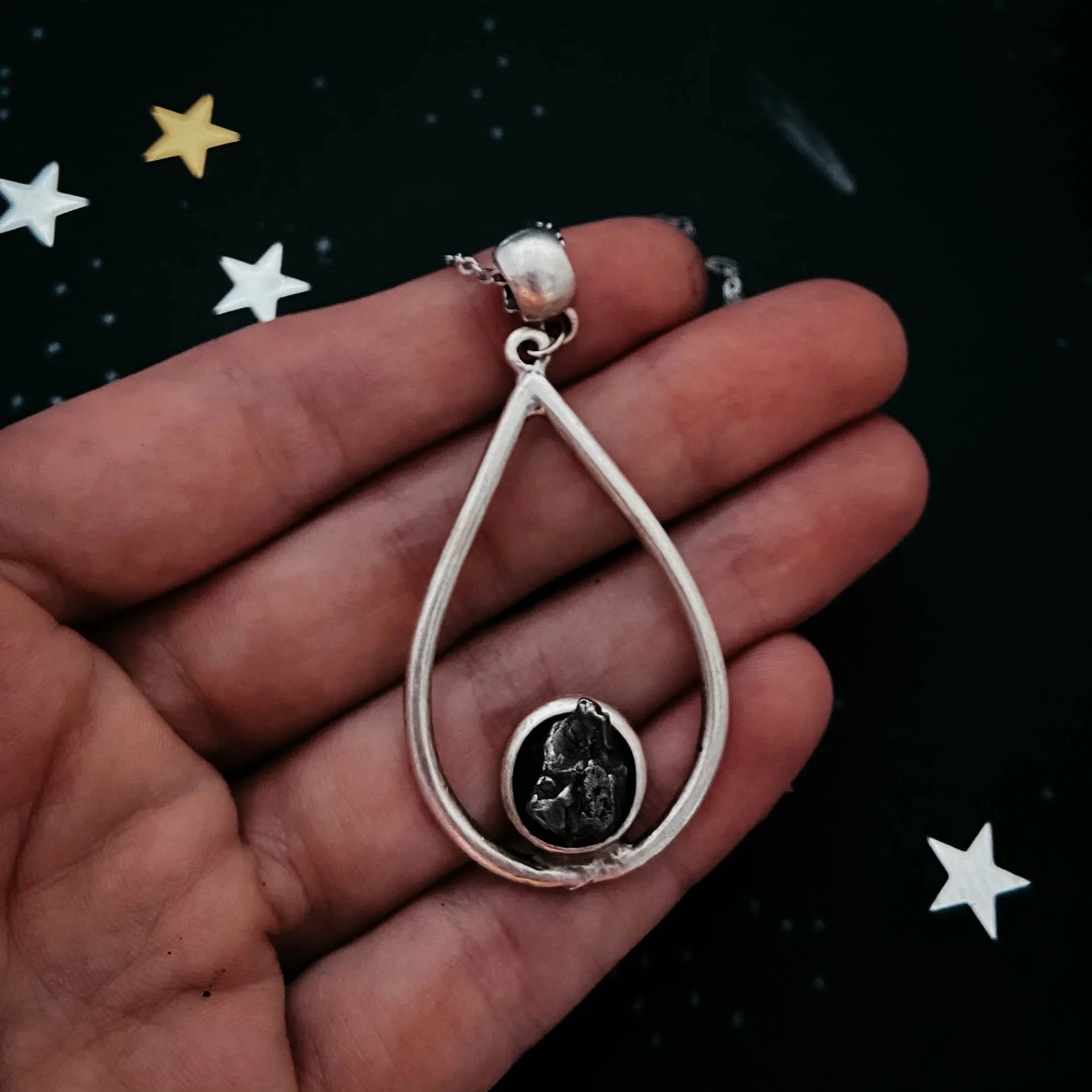 Hand holding silver teardrop pendant necklace with dark irregular stone, starry background