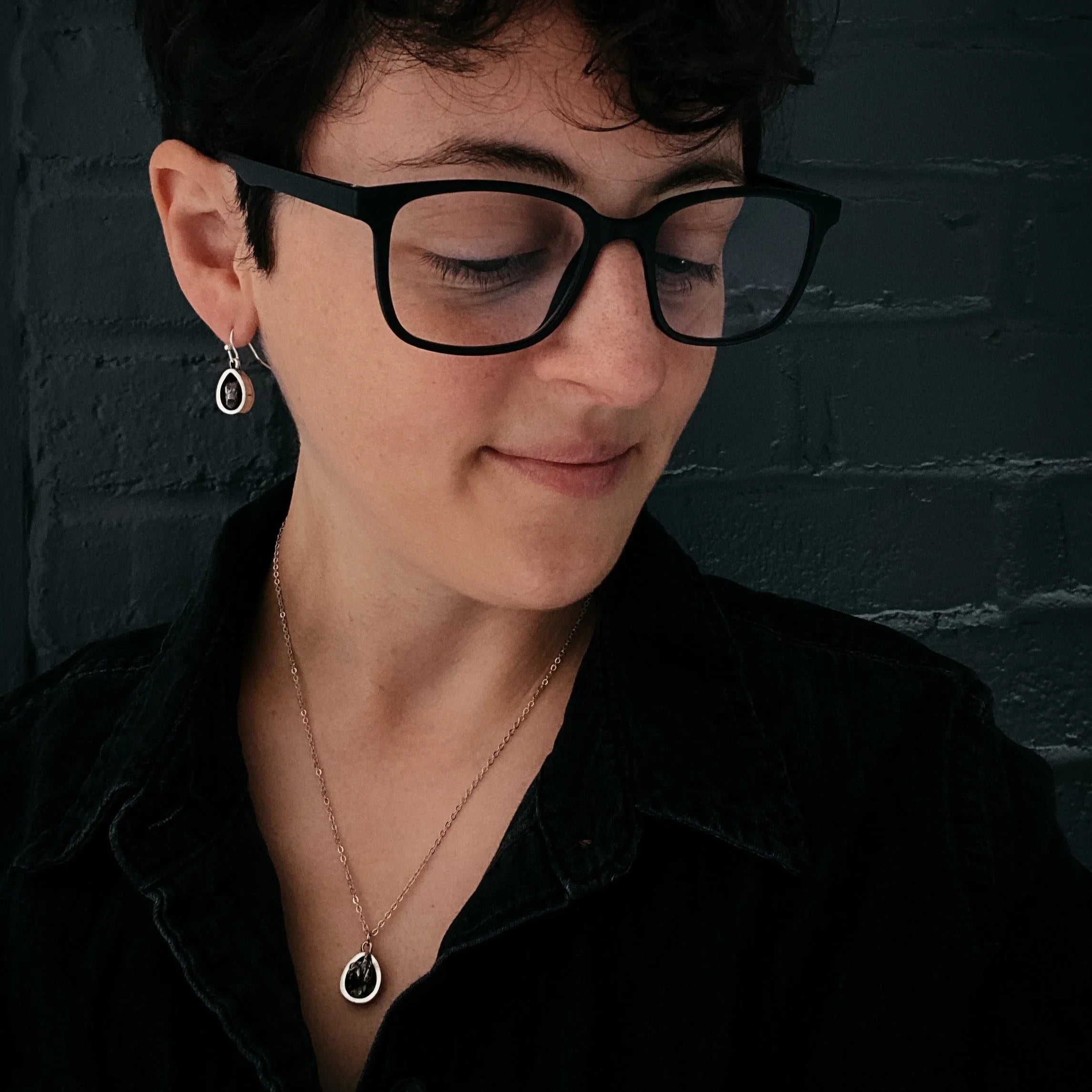 Person with short hair and glasses wearing matching pendant necklace and drop earring, dark top
