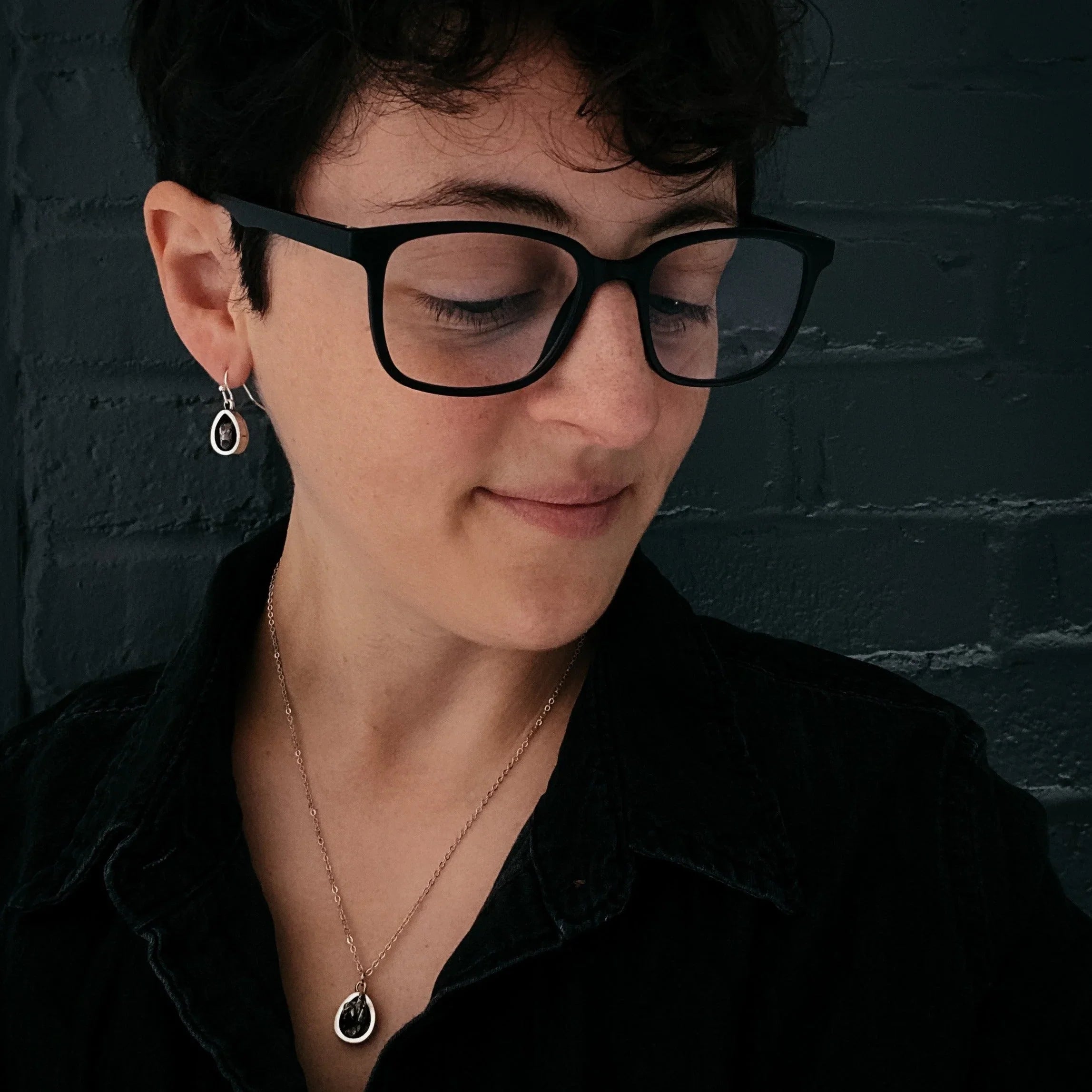 Person wearing black glasses, silver jewelry, and a black shirt against a dark brick wall
