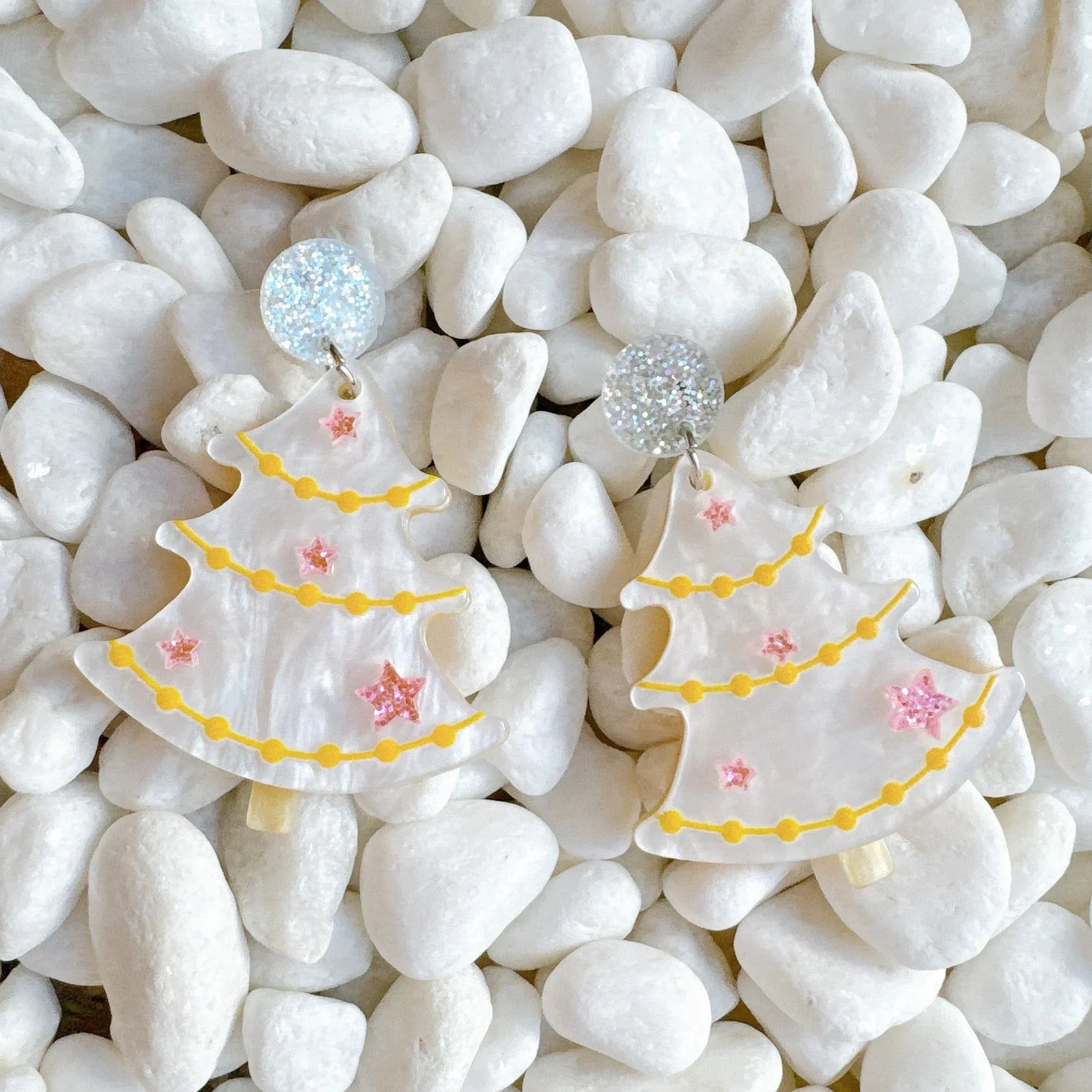 White Christmas tree earrings with yellow garlands and pink stars on white pebbles background