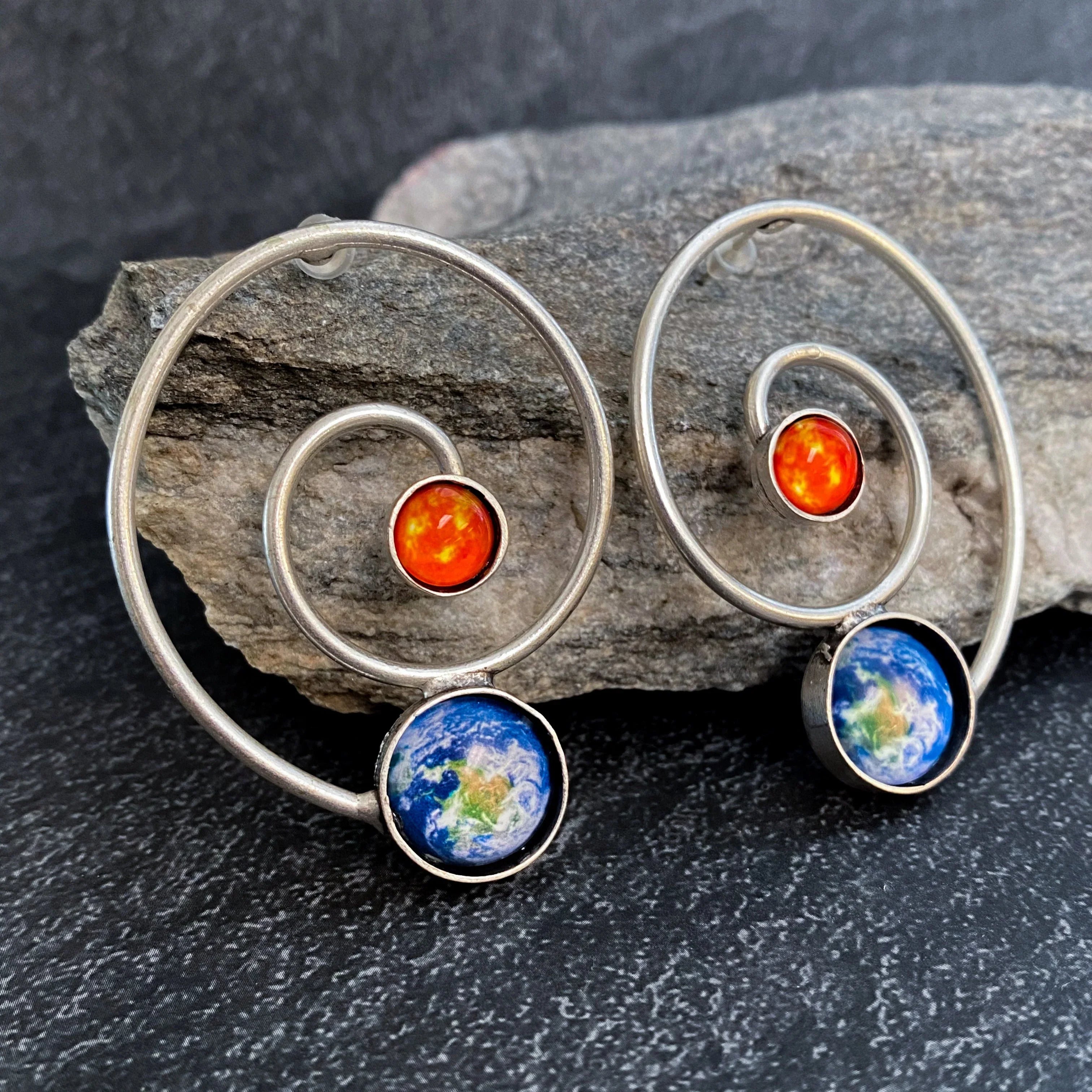 Artist-designed celestial spiral earrings with Earth and sun motifs, displayed on stone