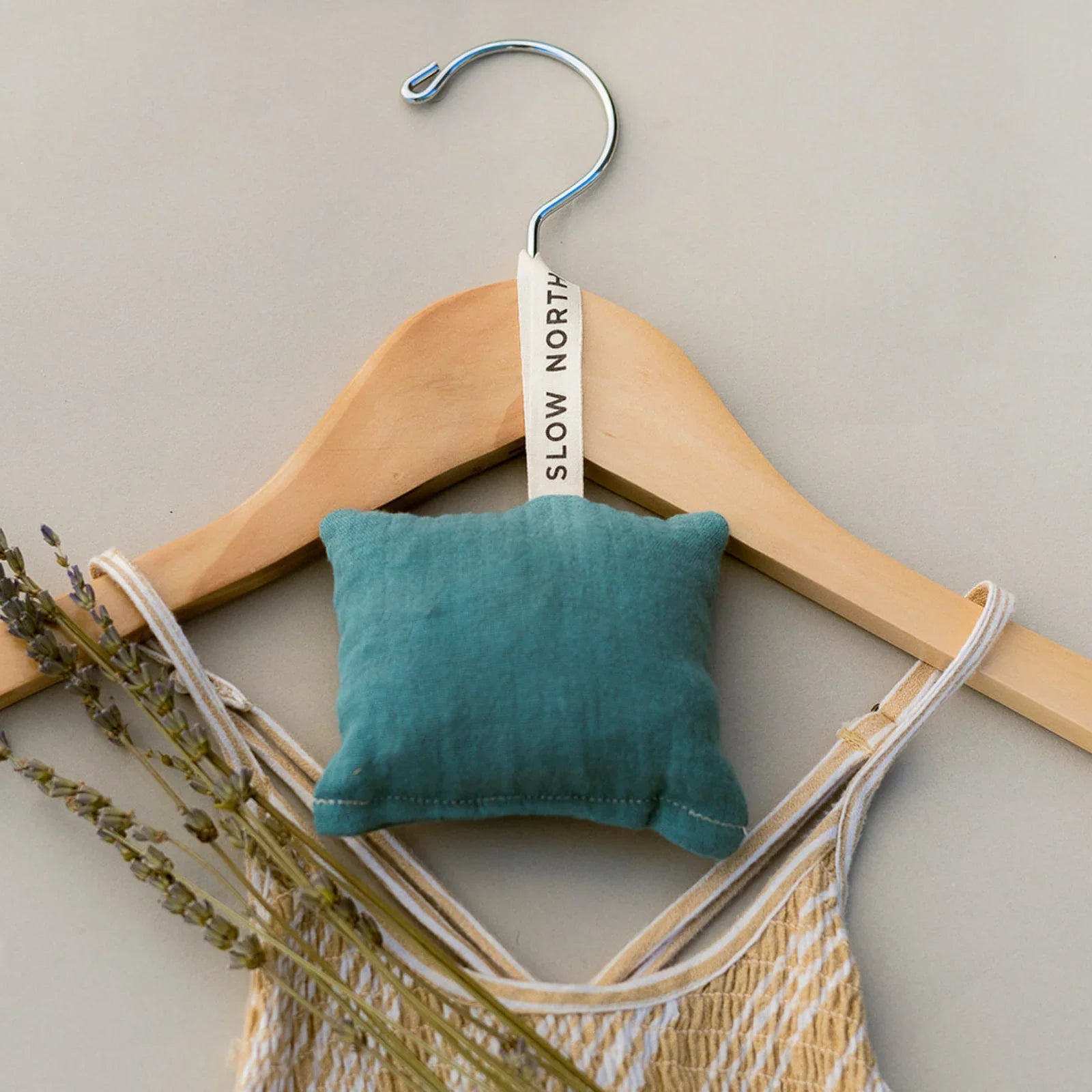 Wooden hanger with a yellow patterned dress, blue hanging sachet labeled Slow North, and dried lavender sprigs