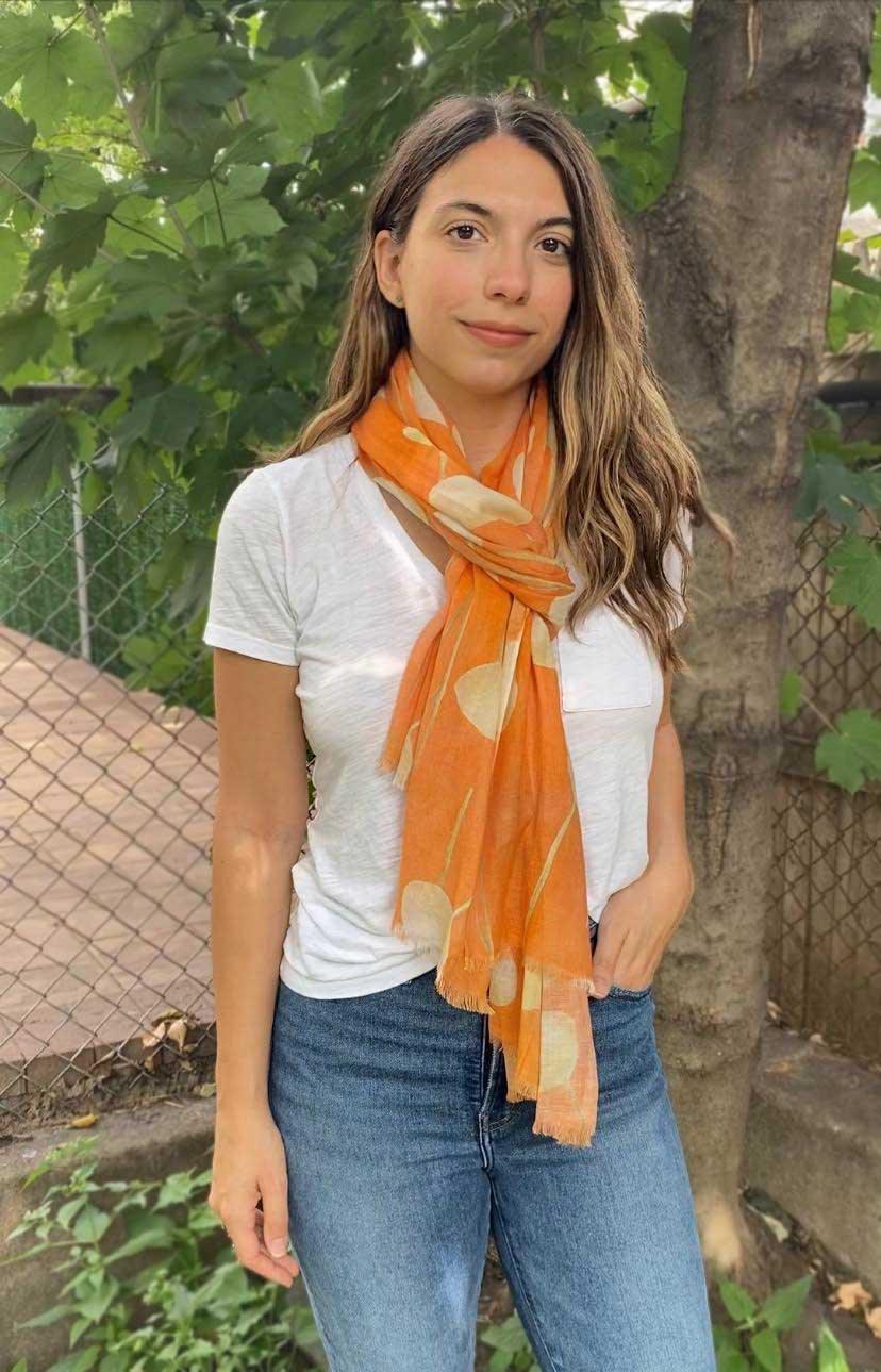 Woman wearing orange floral scarf over white t-shirt and blue jeans standing outdoors near tree and fence