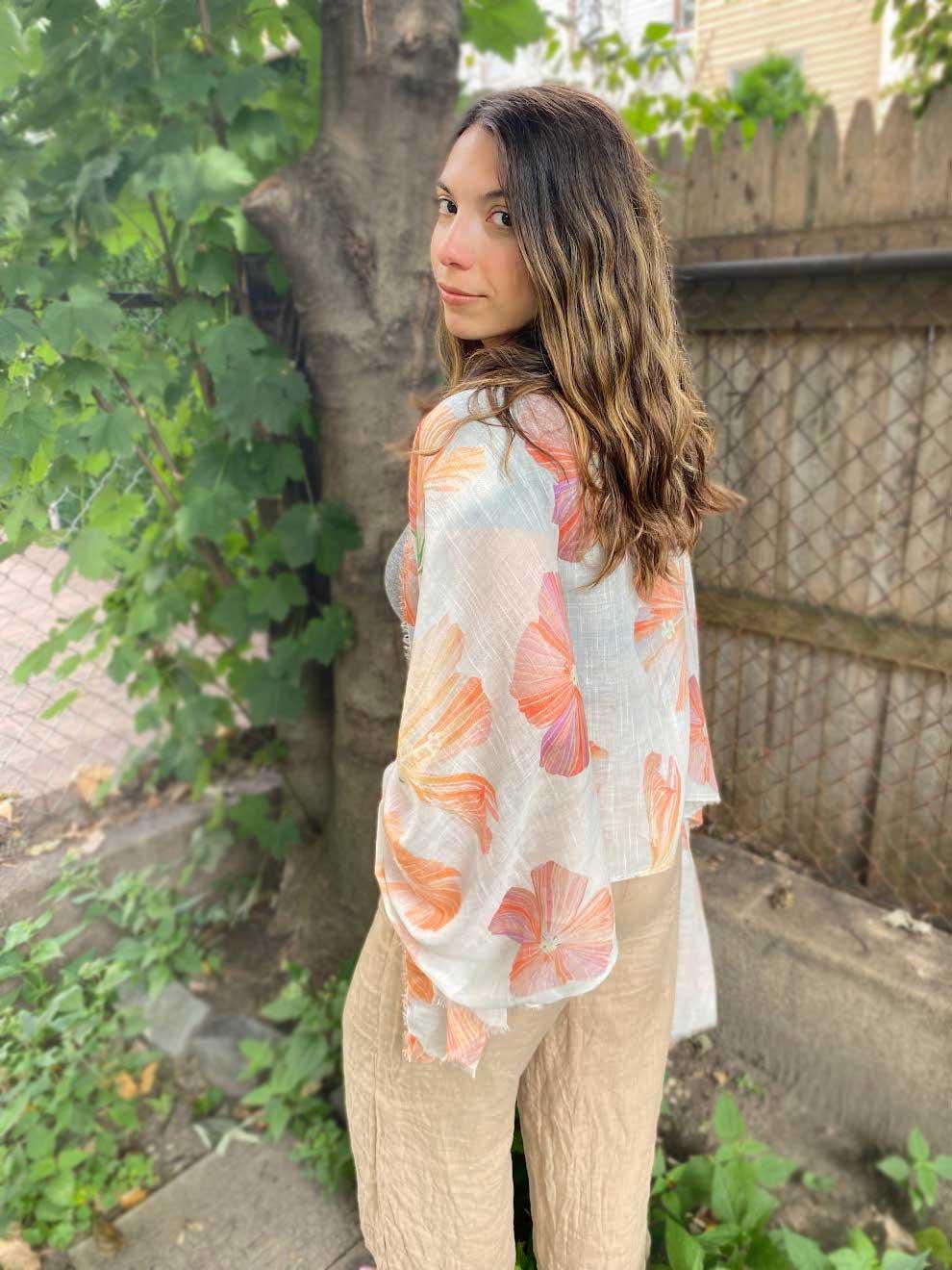Woman outdoors wearing a white scarf with orange floral hibiscus print and beige pants, standing near tree and wooden fence