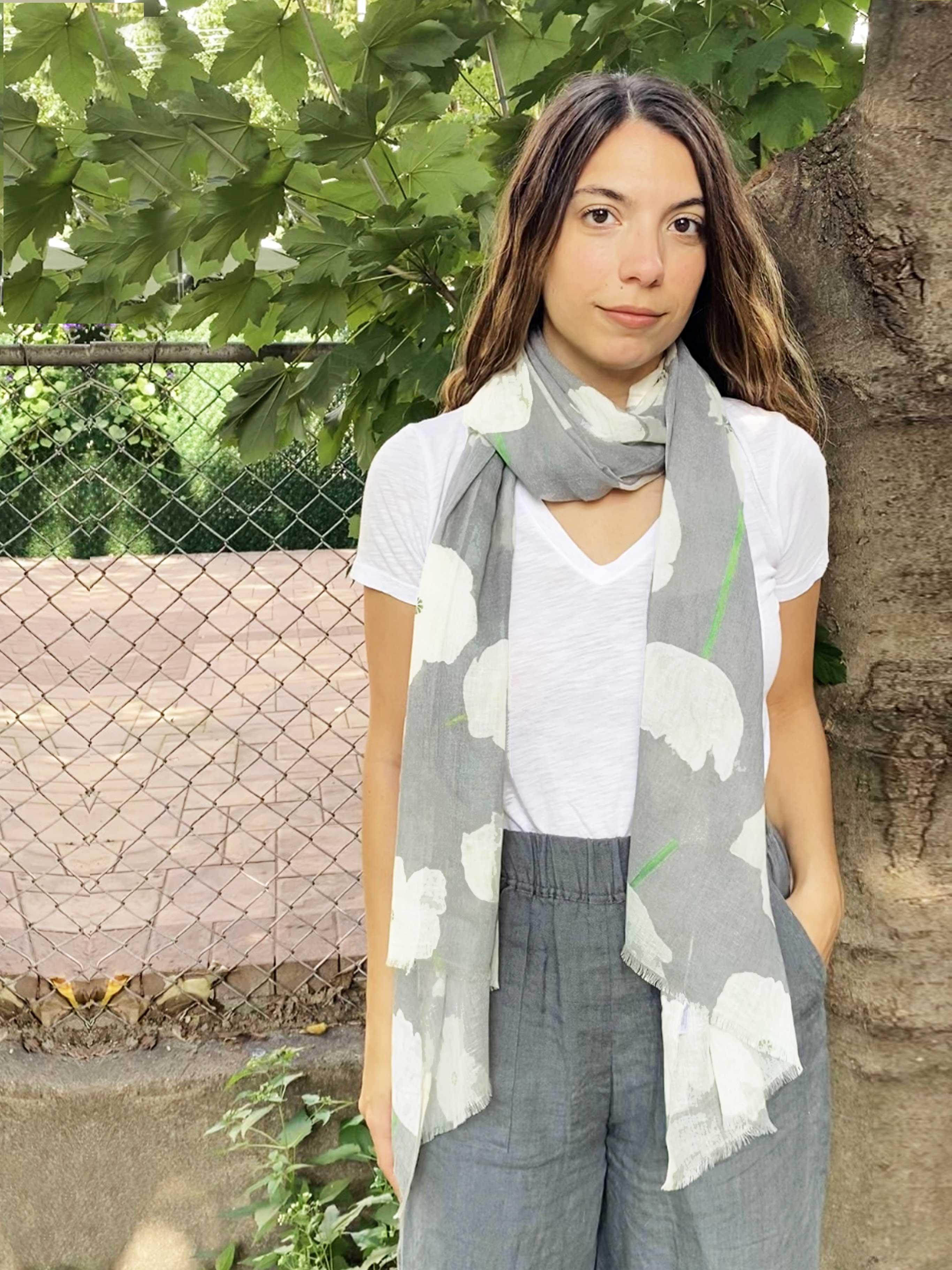 Woman wearing grey floral scarf with white poppies, white t-shirt, and grey pants outdoors next to tree