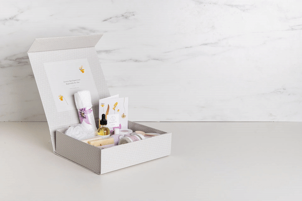 Open white gift box with natural skincare products including soap, oil dropper, towel, and balm on marble surface