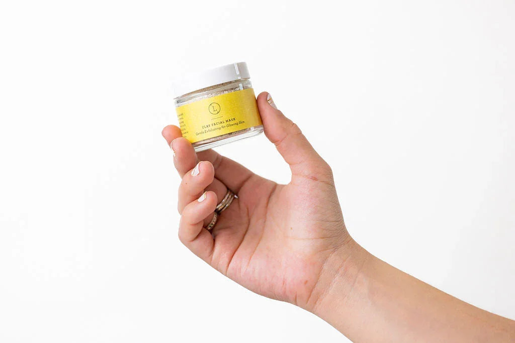 Hand holding small jar of Lizush clay facial mask with yellow label on white background