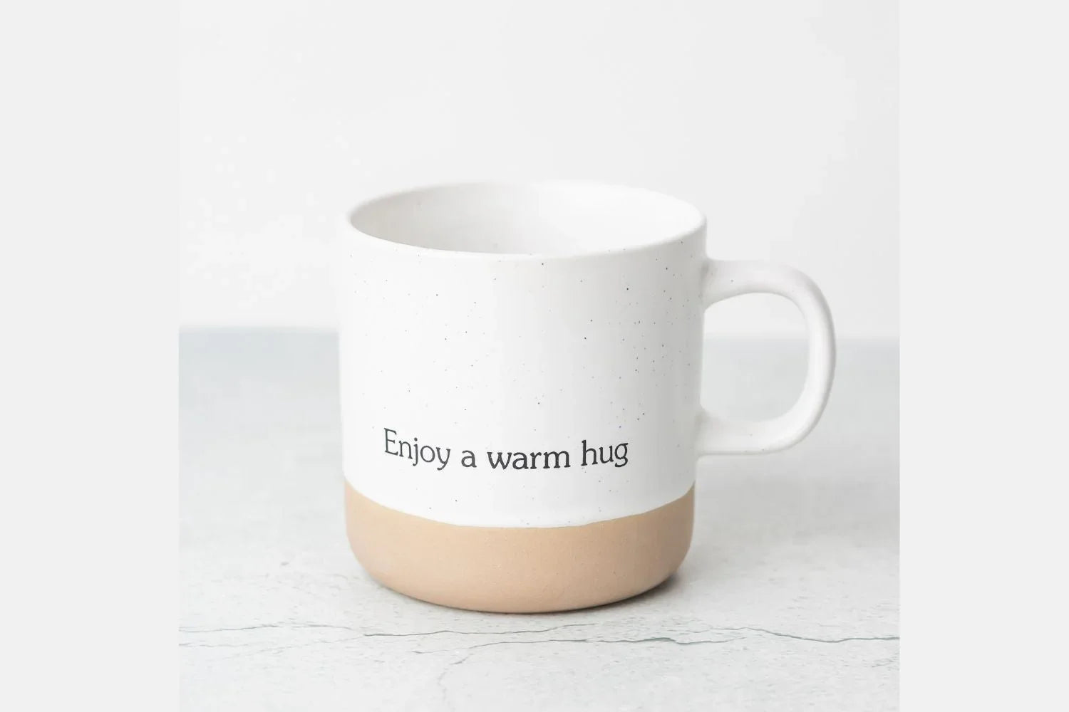 White ceramic mug with beige base and text 'Enjoy a warm hug' on gray surface