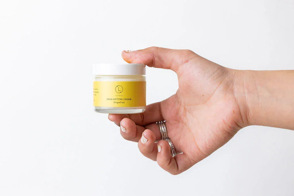 Hand with rings holding a jar of grapefruit shea butter cream with yellow label on white background