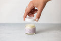 Hand opening jar of lavender scented shea butter cream on gray surface with white background