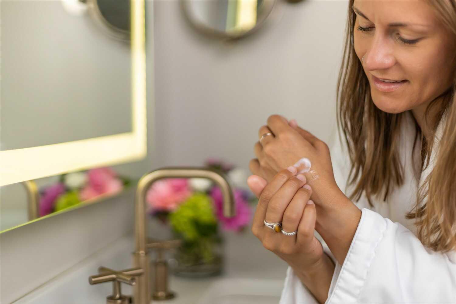 Woman in white robe applying shea butter cream on hand in bright bathroom with flowers