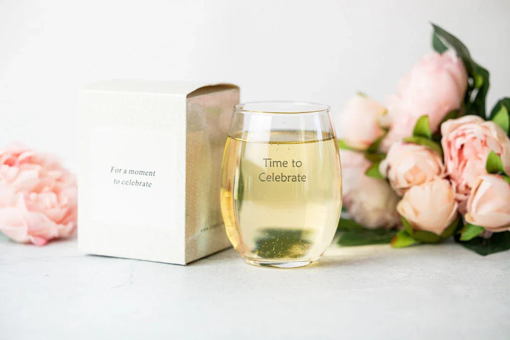 Glass cup filled with sparkling drink labeled Time to Celebrate beside white box and pink peony flowers on light surface