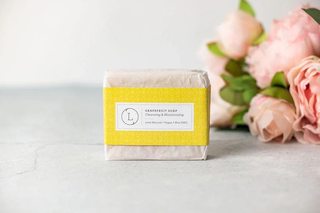 Natural grapefruit soap bar with yellow label on grey surface beside pink flowers