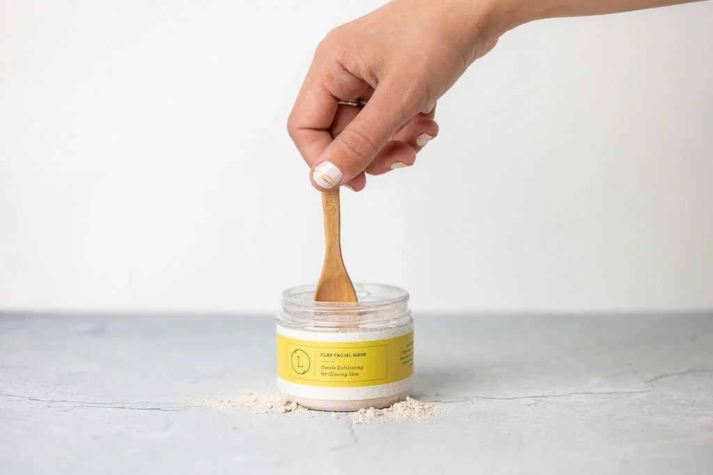 Hand holding wooden spatula dipping into jar of natural clay facial mask with yellow label on gray surface