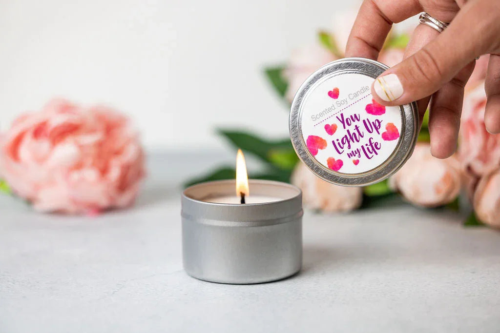 Hand holding tin lid labeled You Light Up My Life above lit scented soy candle with pink flowers in background