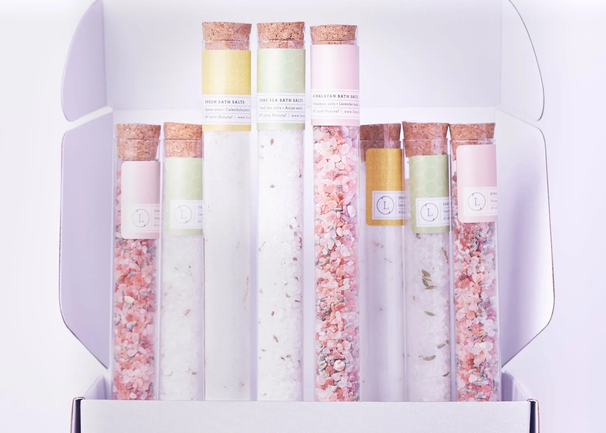 Set of glass test tubes with cork stoppers containing pink and white bath salts in a white open box