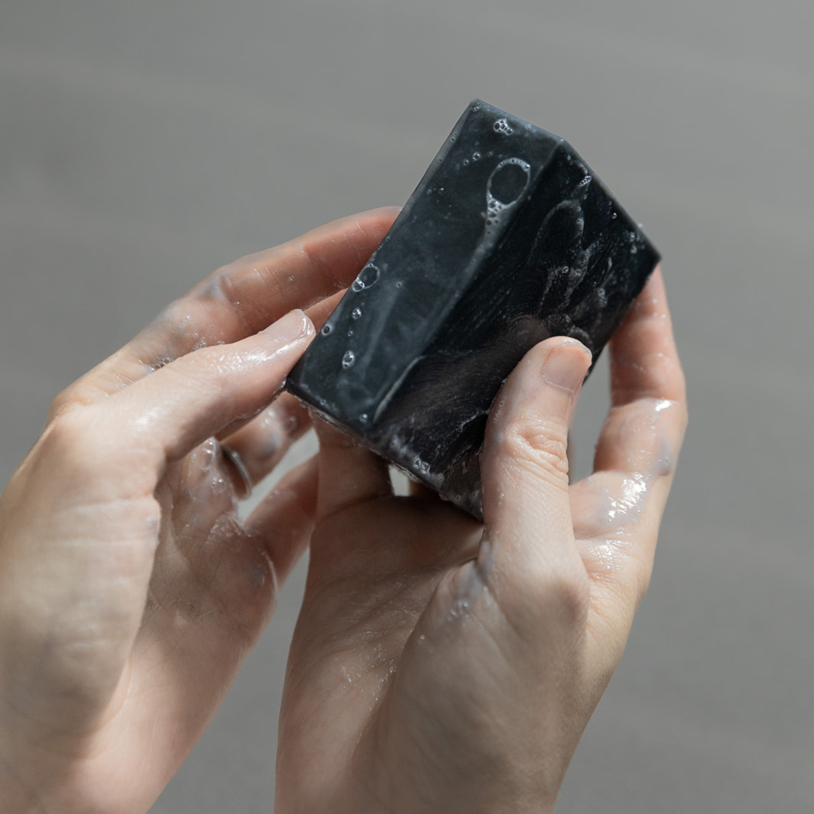 Wet hands holding a black bar of soap with bubbles on a gray background