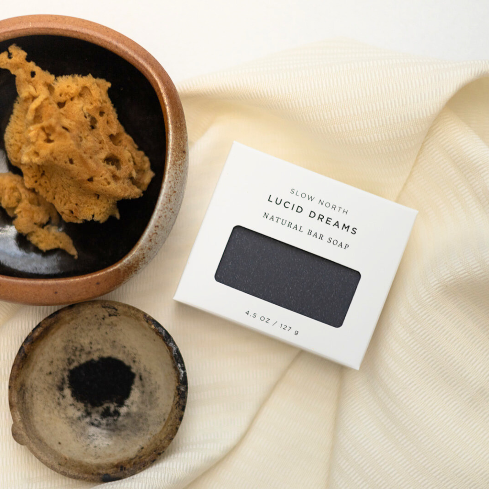 Slow North Lucid Dreams natural bar soap packaging with black soap bar on cream fabric next to ceramic bowls