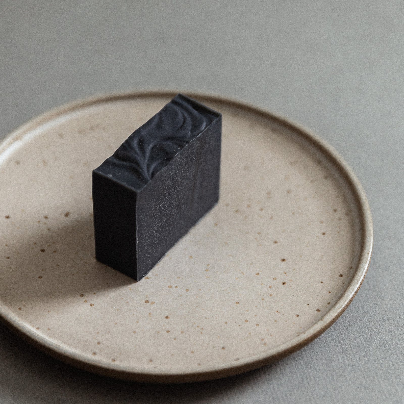 Black natural bar soap with textured top on beige ceramic plate on gray surface