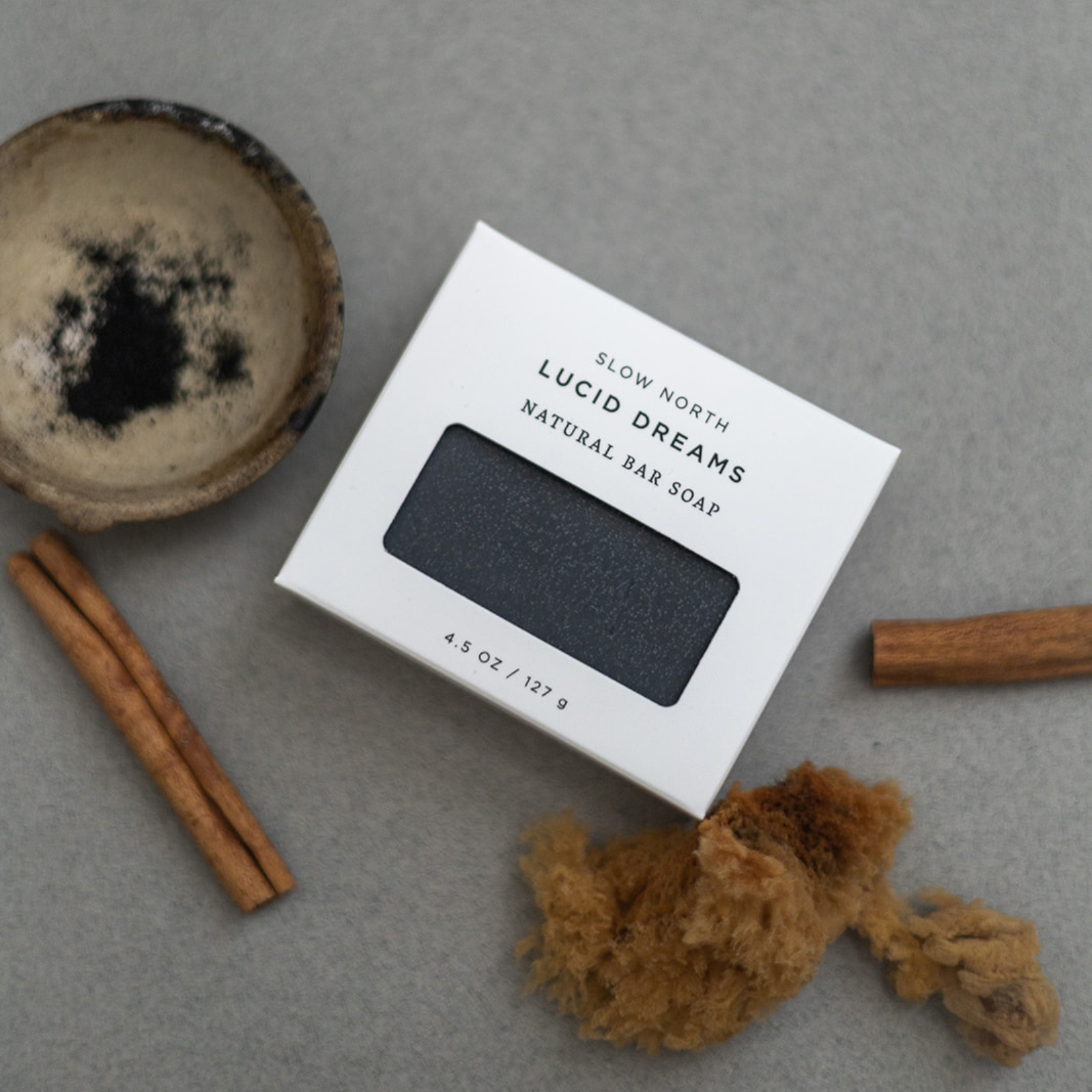 Lucid Dreams natural bar soap by Slow North in white box with cinnamon sticks, black powder in bowl, and brown sponge