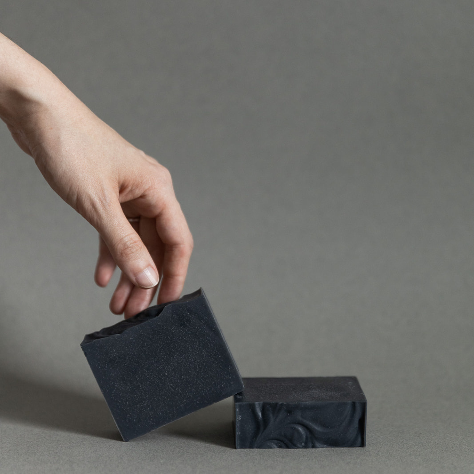 Hand holding handcrafted black natural soap bar next to another soap bar on grey background