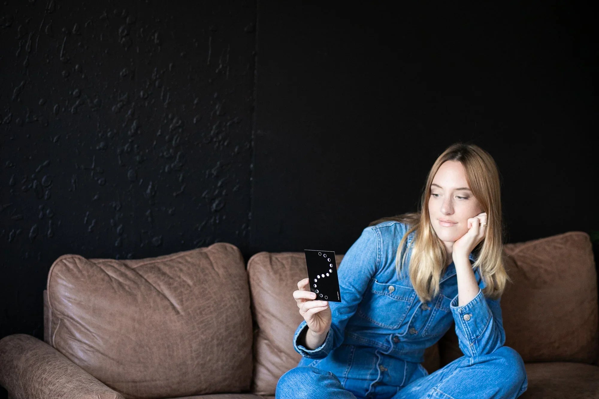 Woman in denim outfit sitting on brown leather couch holding a black card with lunar phases on black wall background