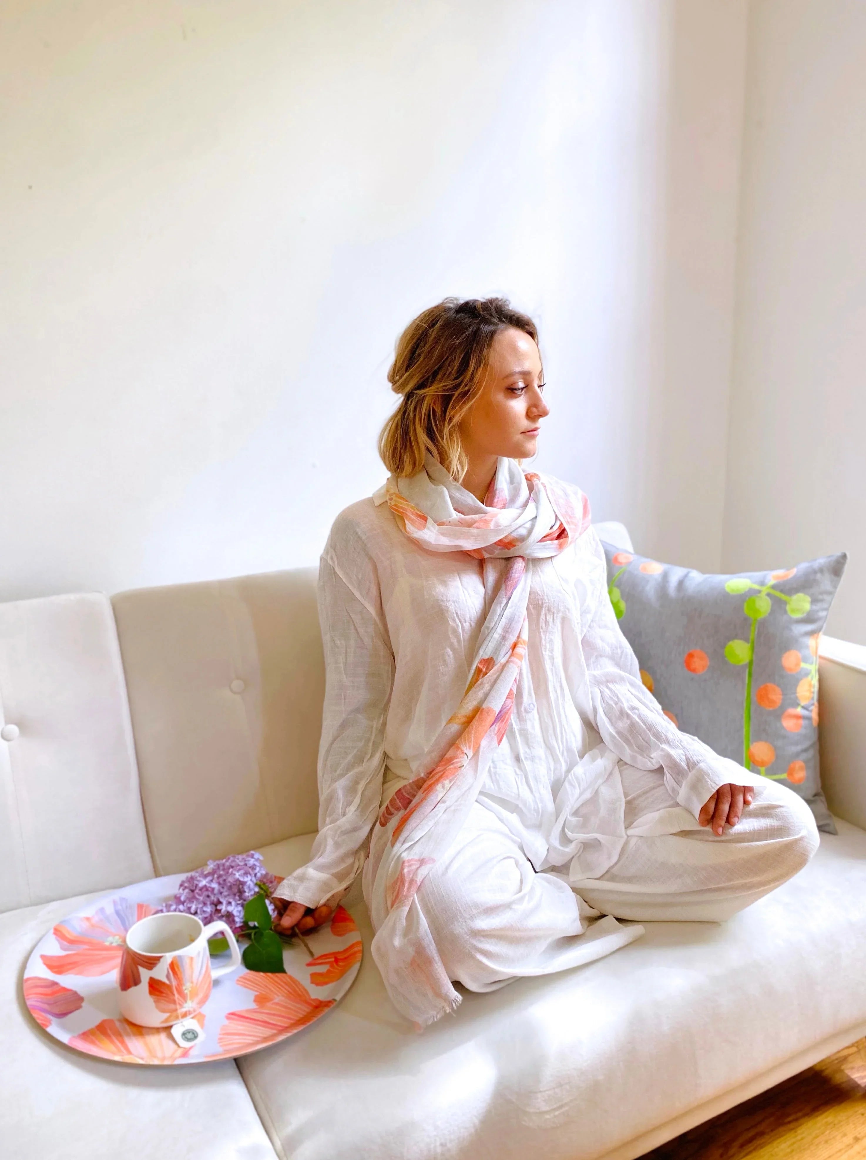 Woman in white outfit and floral scarf sitting cross-legged on white couch with decorative pillow