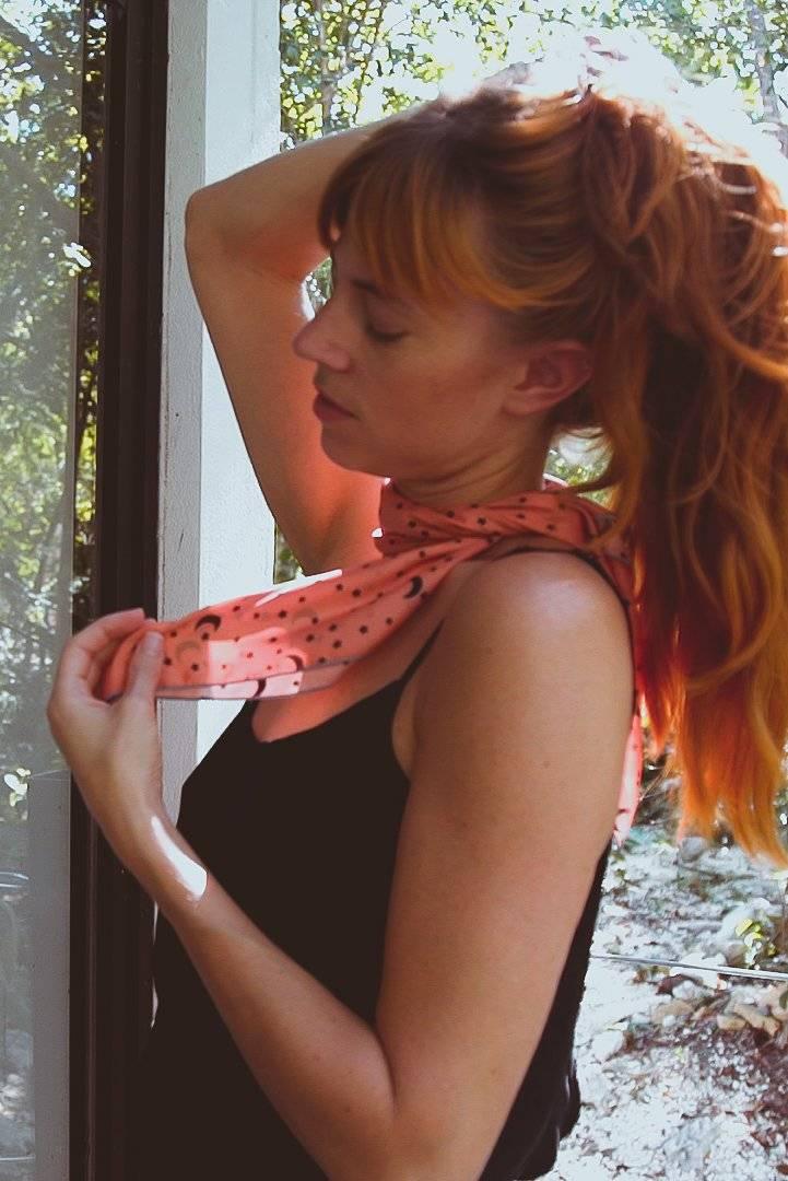 Red-haired woman in black tank top adjusting pink patterned neck scarf near window with outdoor greenery