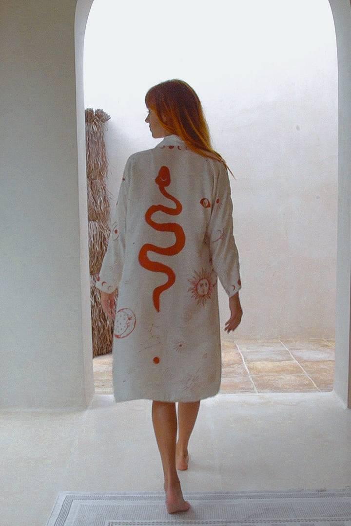 Woman walking barefoot in a light room wearing a white robe with a large red snake and sun design on the back