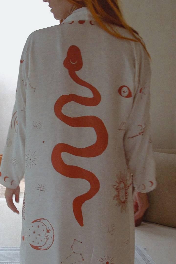 Back view of woman wearing white robe with large red snake and celestial prints in indoor setting