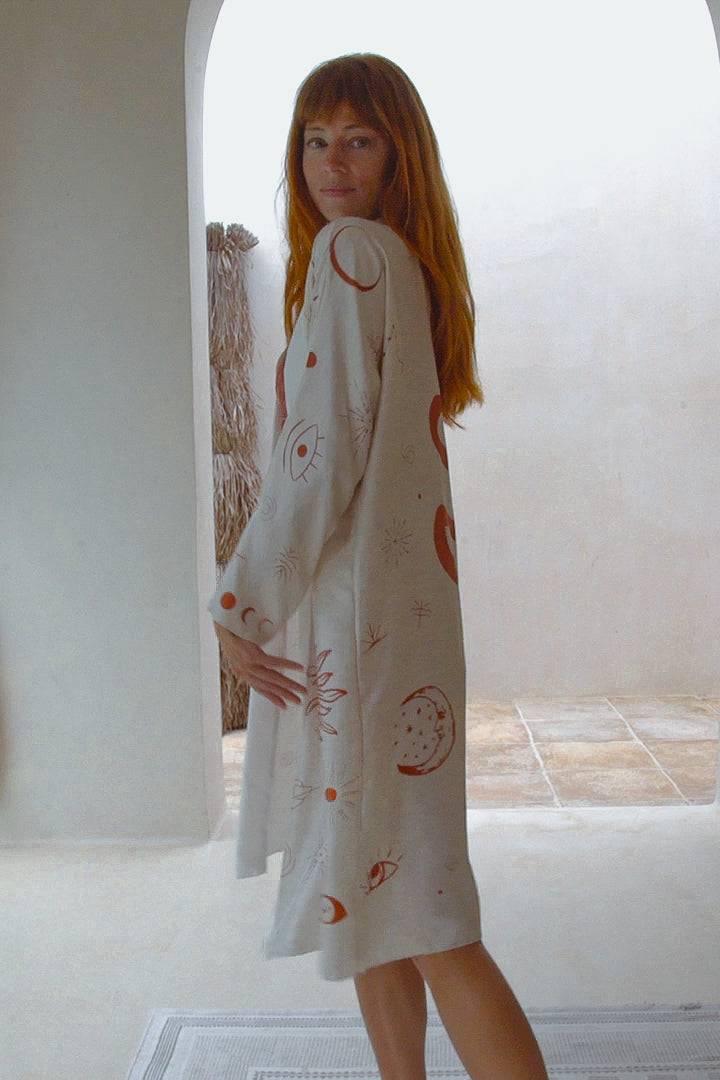 Red-haired woman wearing a beige dress with abstract sun, moon, and eye prints in a bright minimalist room