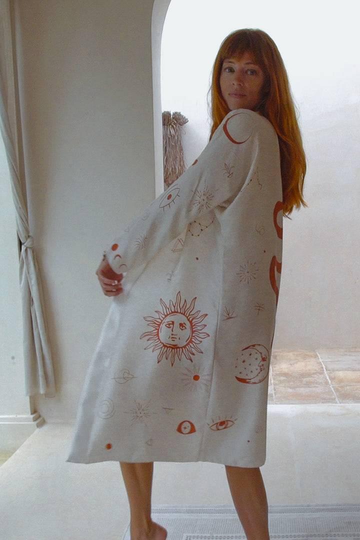 Woman wearing light robe with sun, moon, and eye celestial prints in bright minimal room