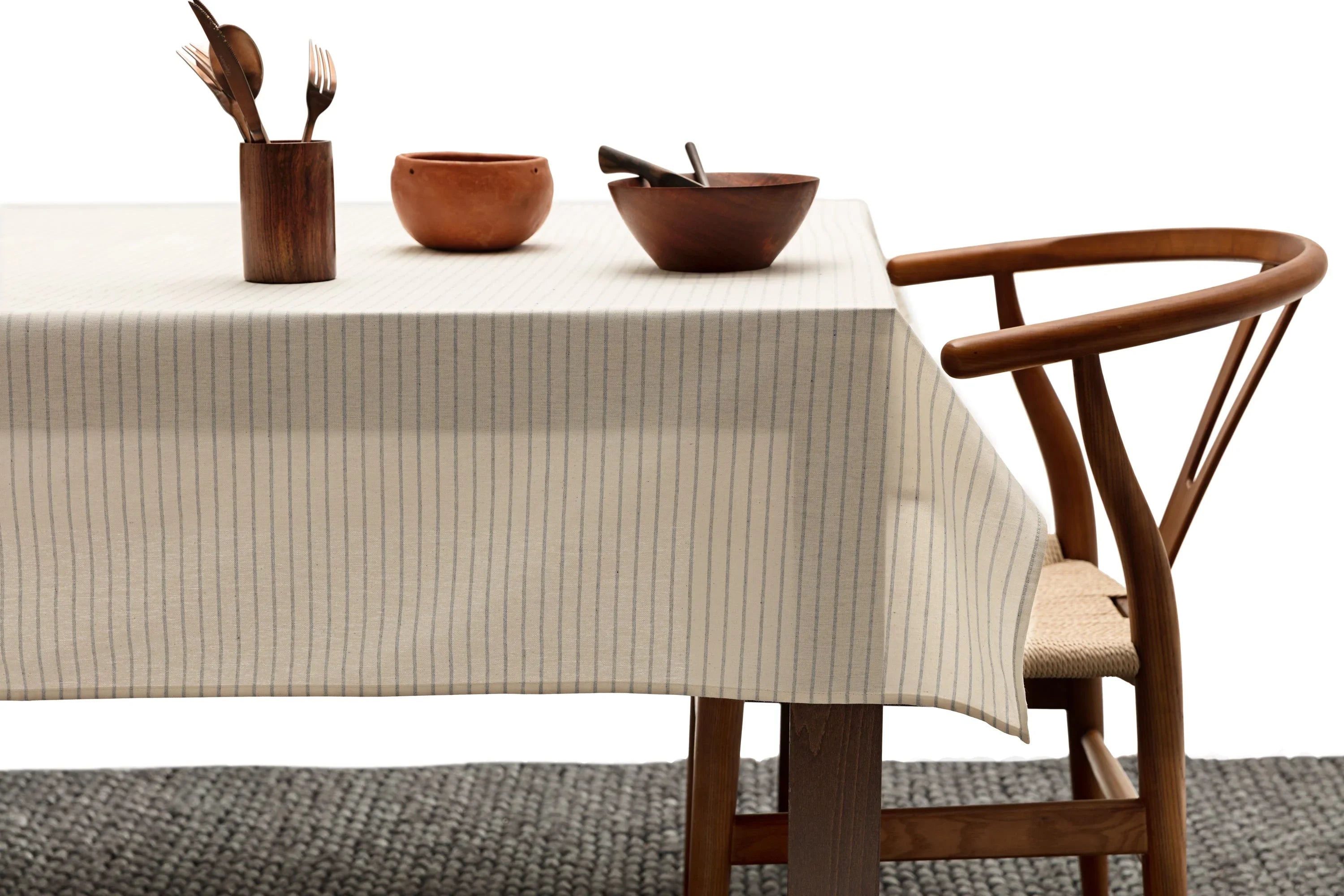 Neutral striped tablecloth on dining table with wooden bowls, utensils, and modern wood chair