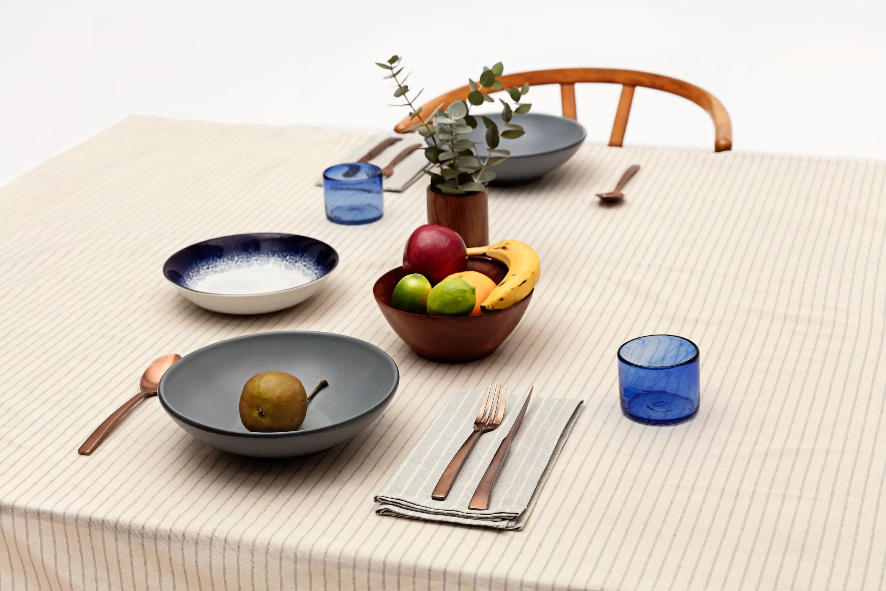 Minimalist table setting with striped tablecloth, blue glasses, fruit bowl, and modern dishware at Qu!rk Boutique.