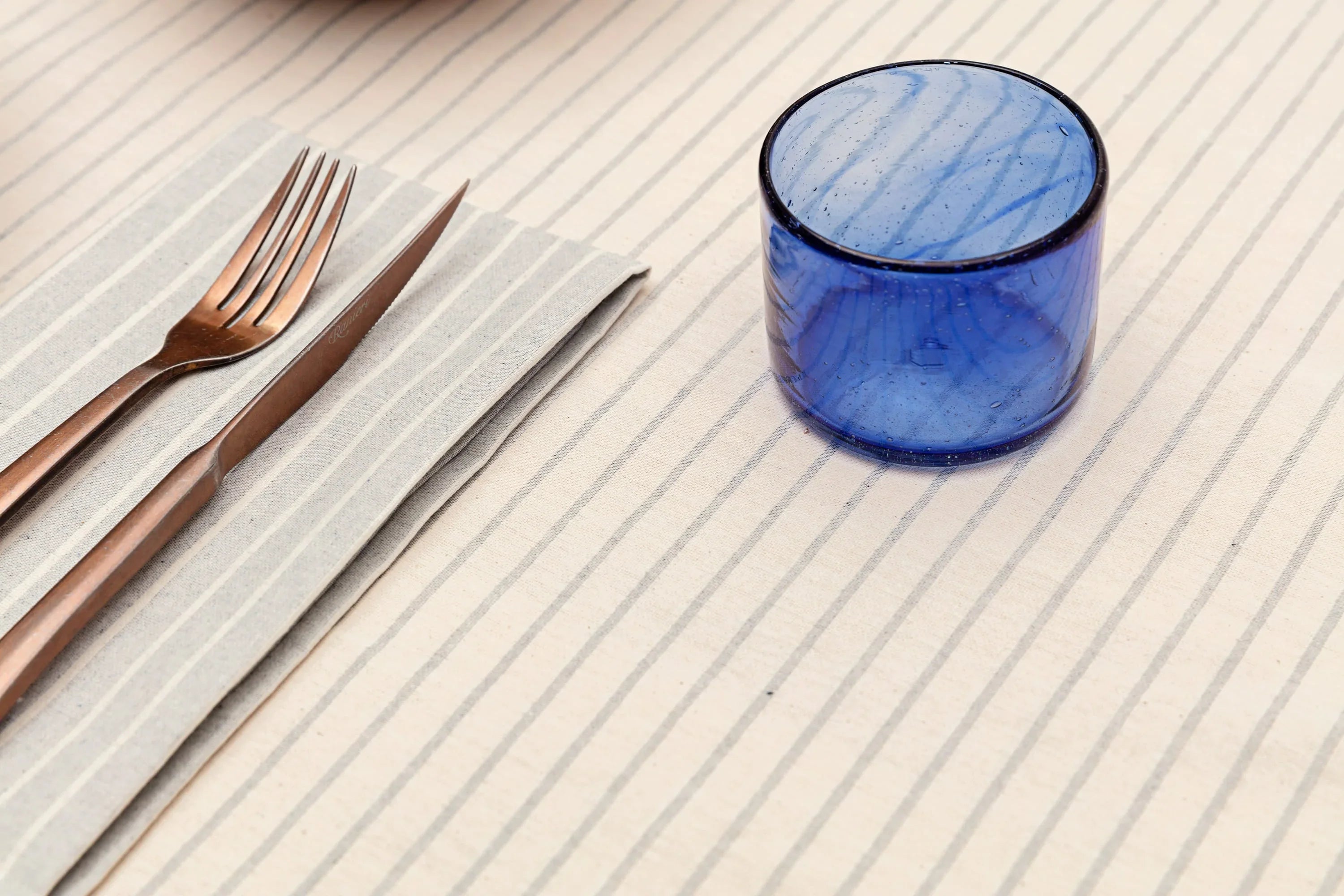 Blue glass cup, bronze cutlery on striped cloth napkin, minimalist table setting, Qu!rk Boutique