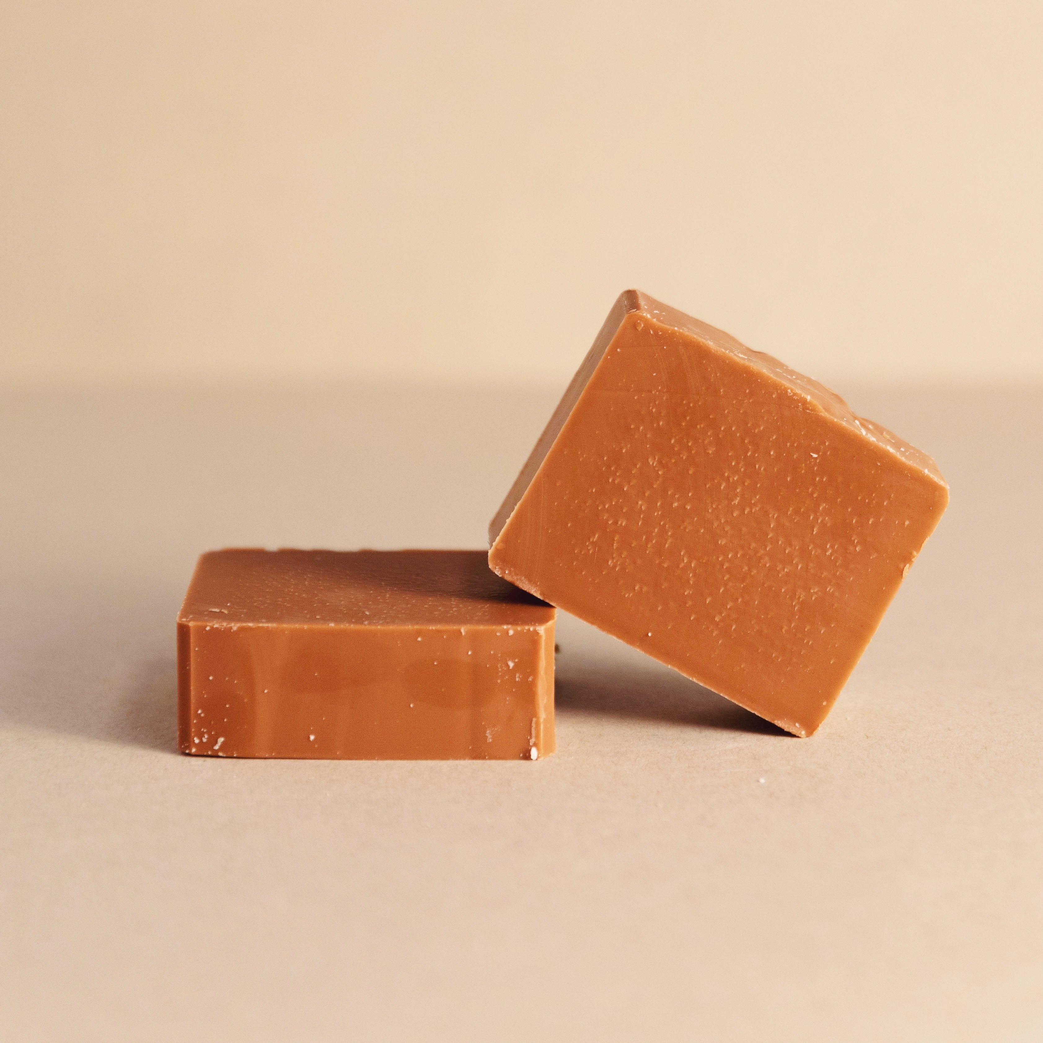 Two smooth caramel fudge squares on a beige surface with warm lighting