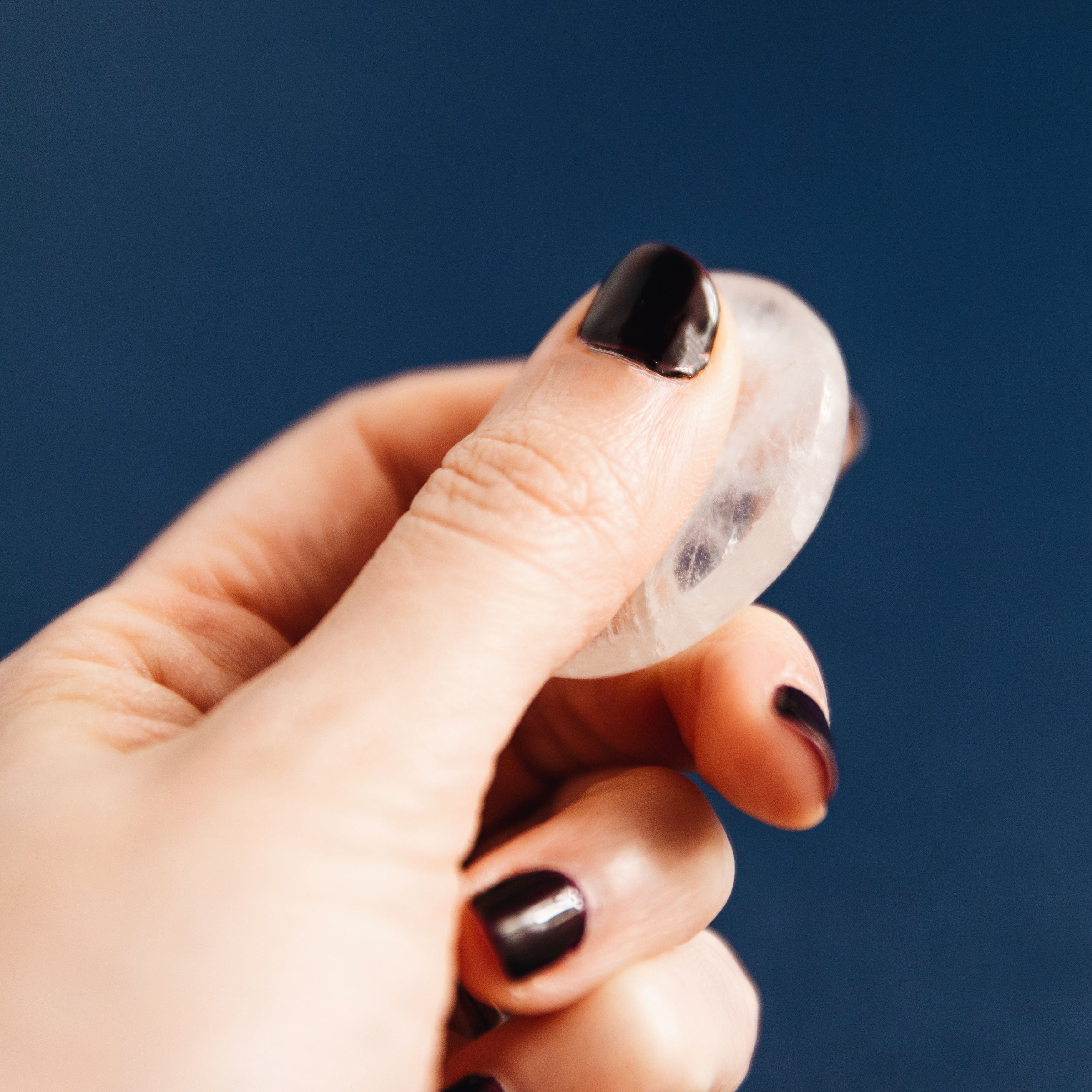 Hand with black nail polish holding a clear polished crystal against a dark blue background