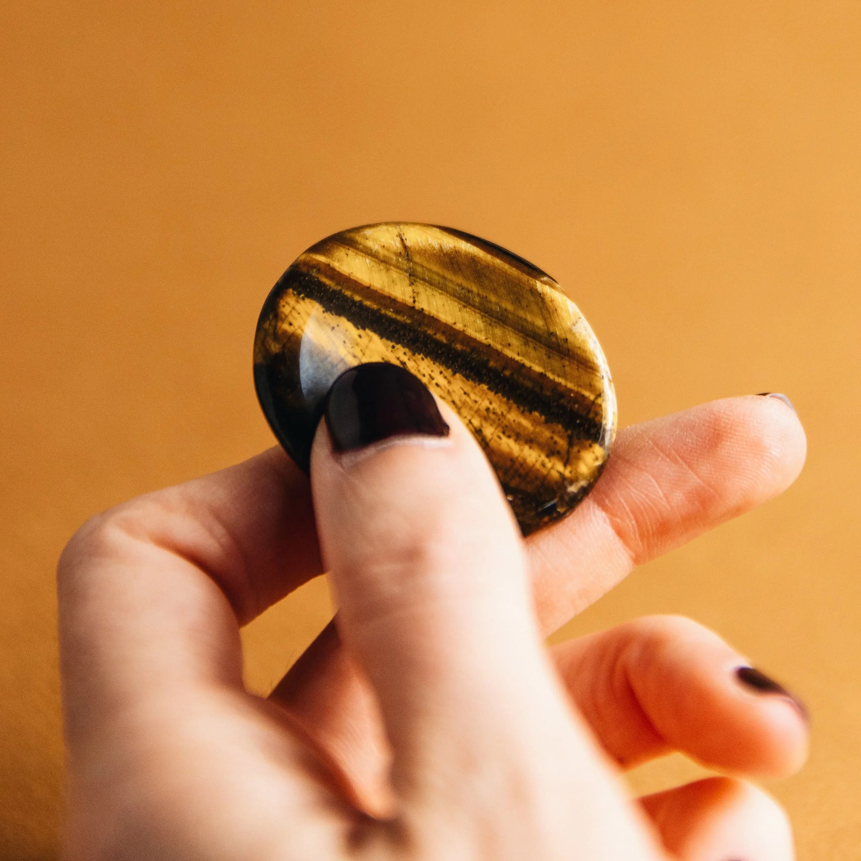 Hand holding polished tiger's eye stone with brown and golden stripes on orange background