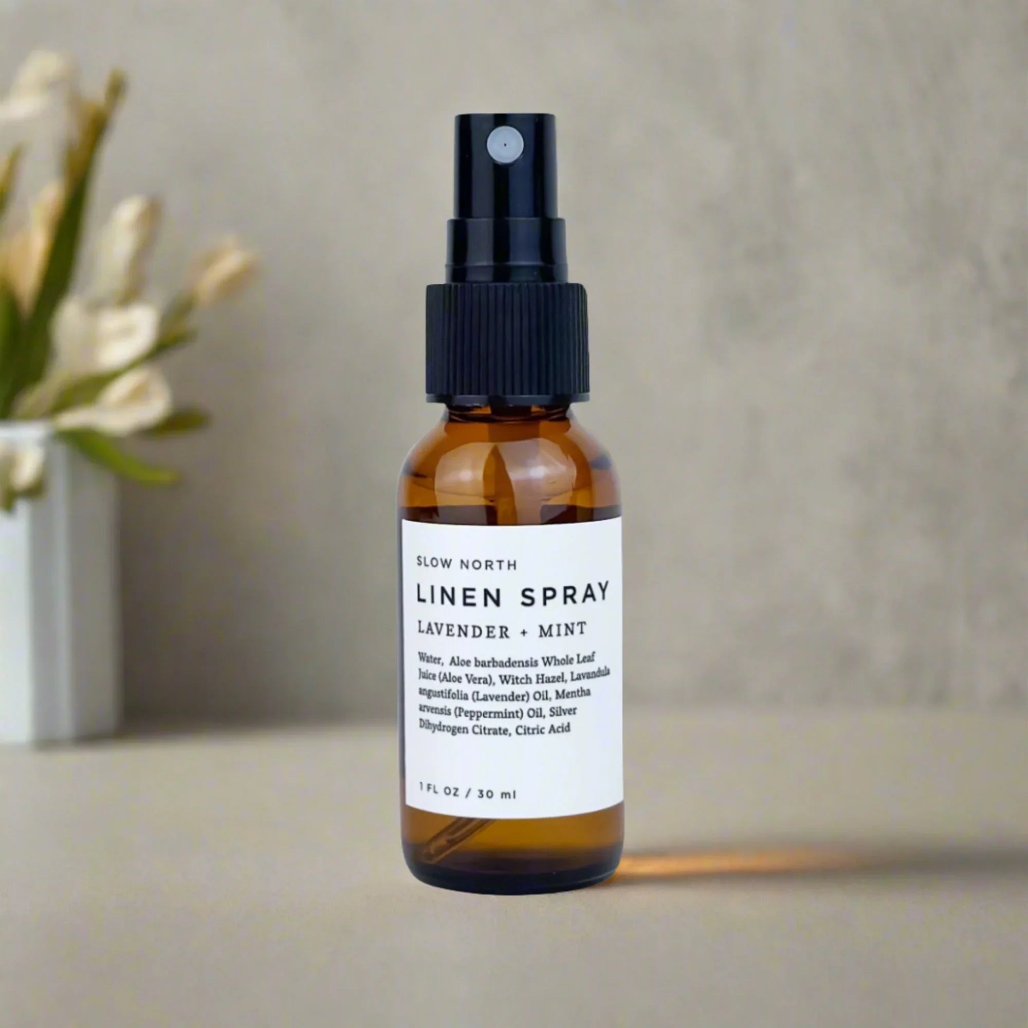 Amber glass bottle of Slow North Lavender + Mint linen spray on neutral surface with blurred flowers in background