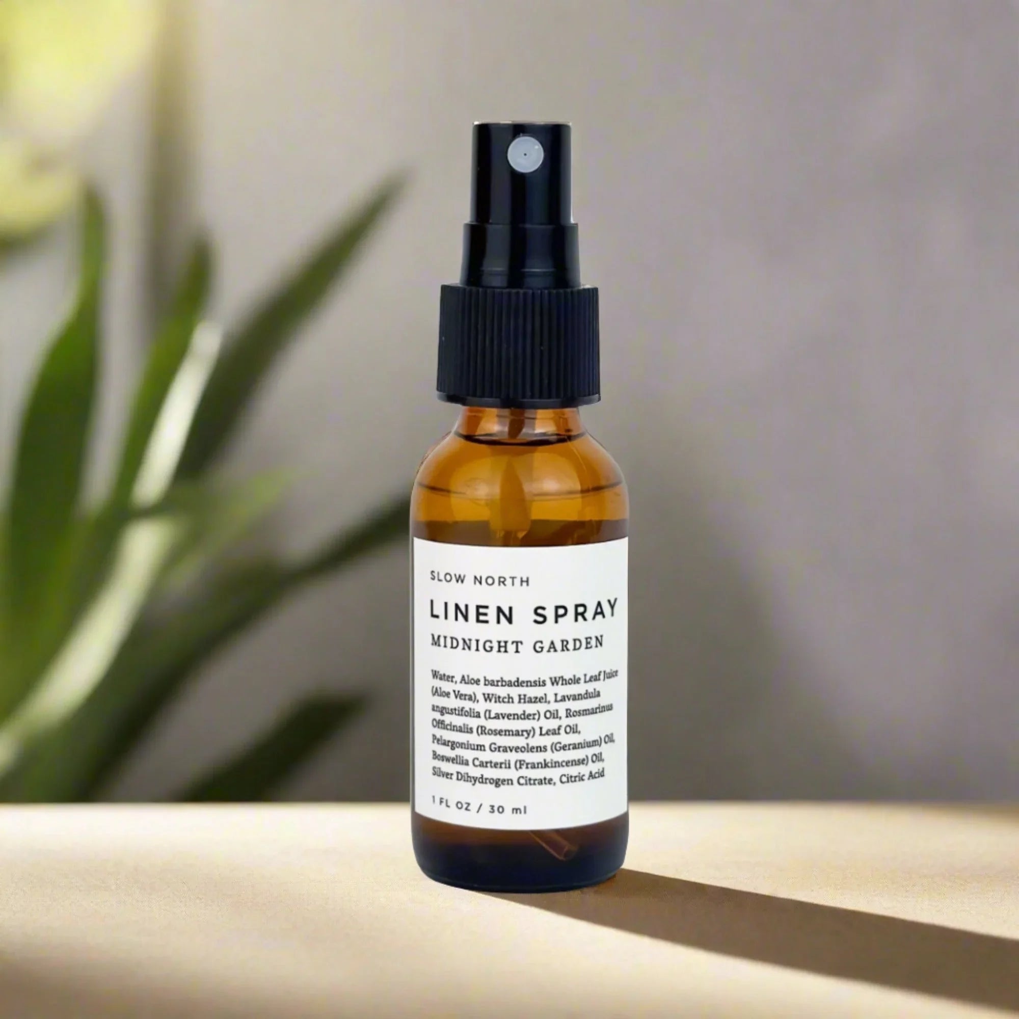 Amber glass bottle of Slow North Midnight Garden linen spray on beige surface with blurred green plant