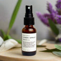 Slow North Shangri-La linen spray in amber bottle with spray top on wooden surface with blurred purple flowers