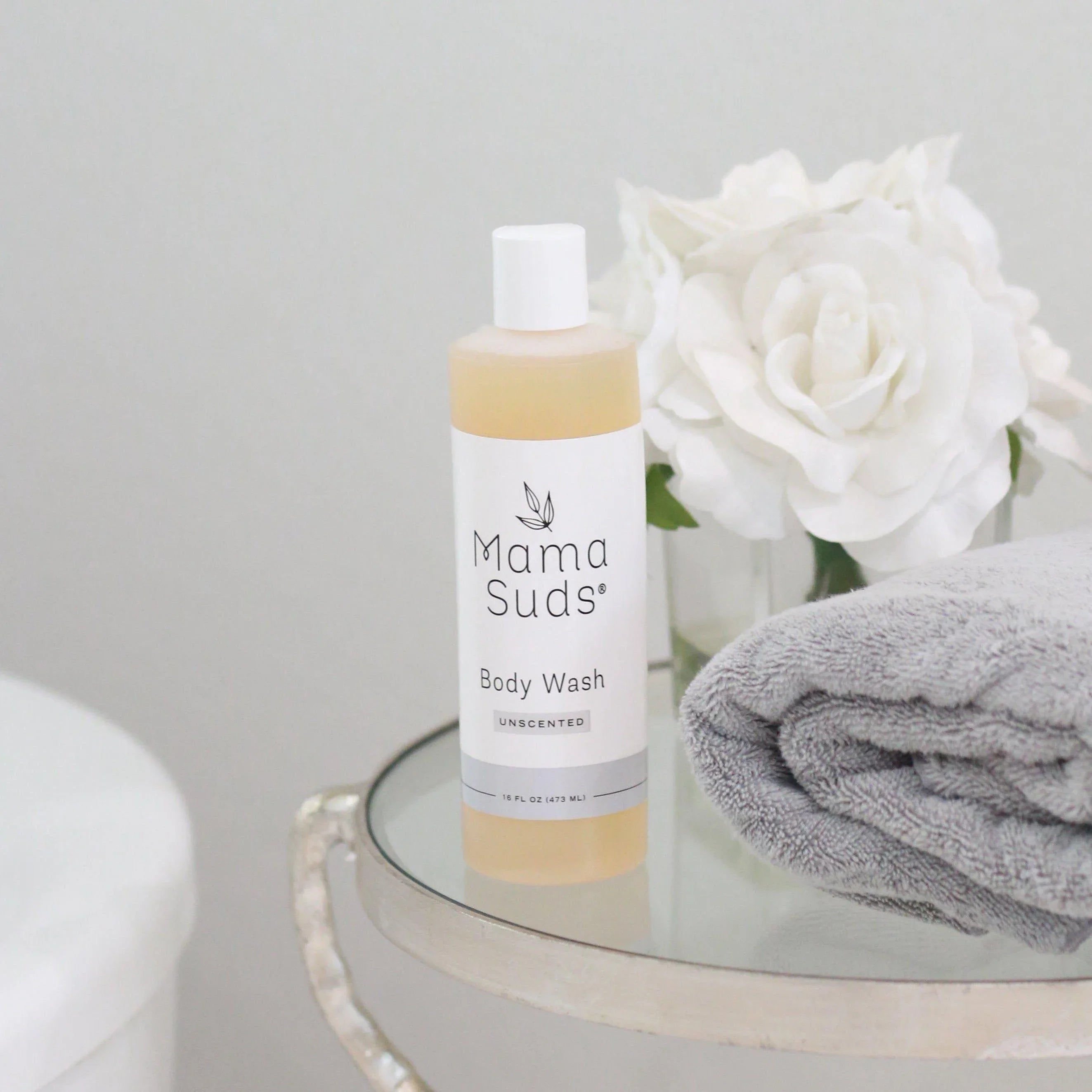 Mama Suds unscented body wash on glass table with gray towel and white roses, boutique spa setting