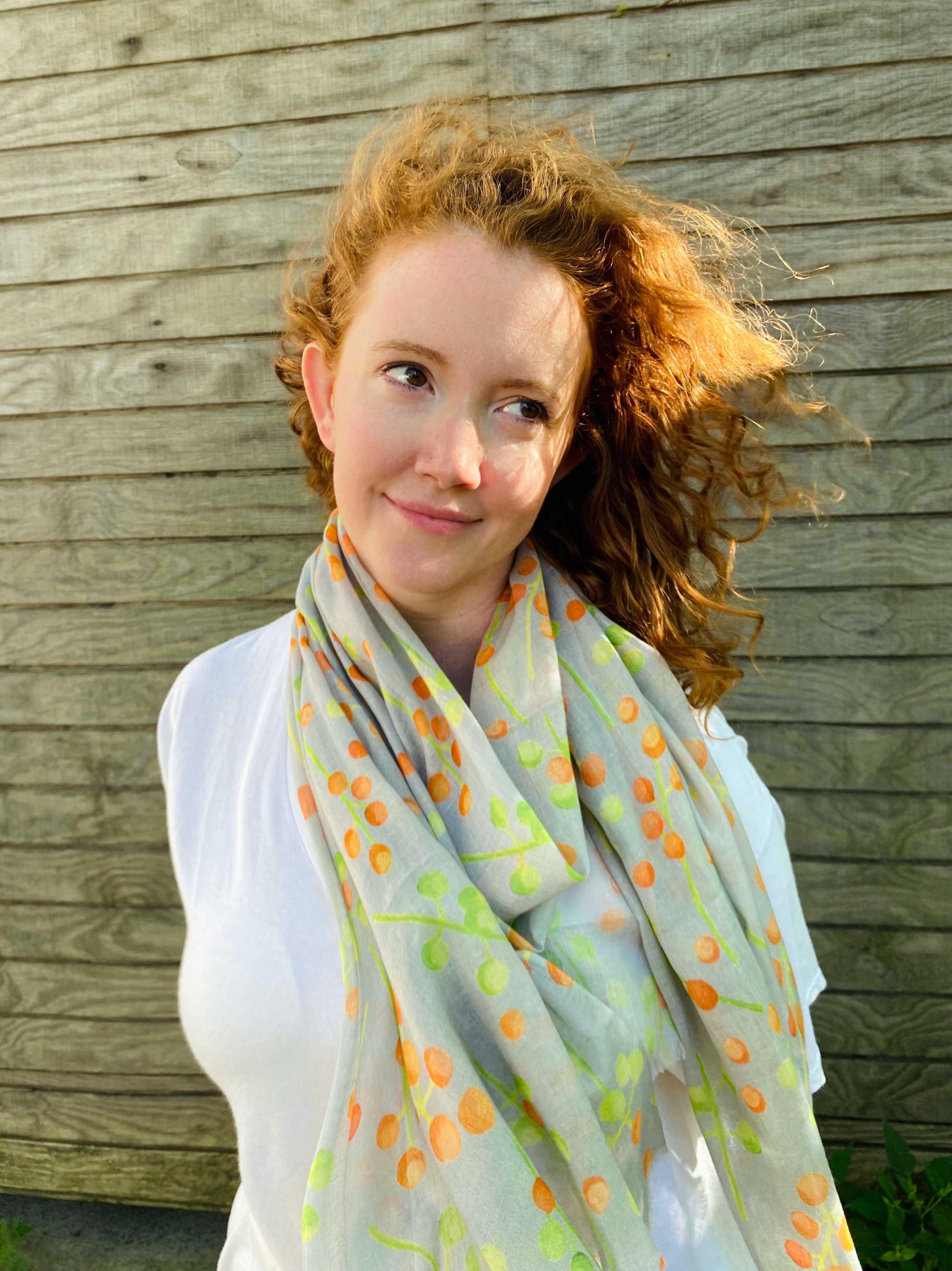 Red-haired woman wearing a white shirt and colorful polka dot scarf standing in front of wooden wall