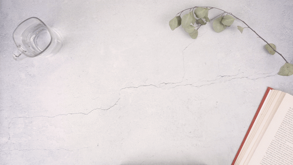 Natural skincare oil being poured onto a hand with eucalyptus branch, glass of water, and open book on white surface